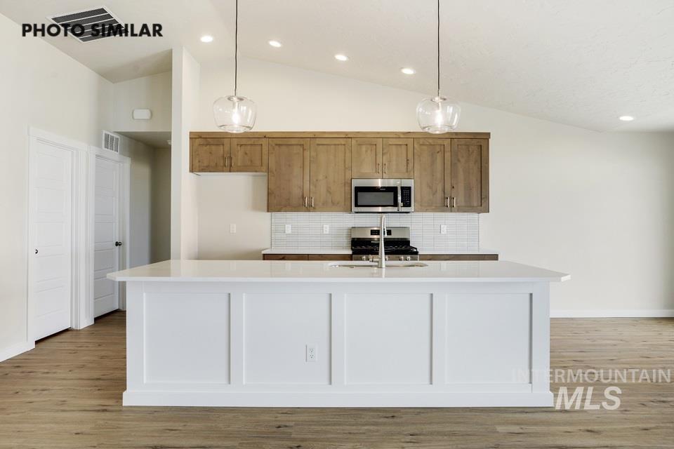 Kitchen with tasteful backsplash, hanging light fixtures, appliances with stainless steel finishes, light wood-type flooring, and an island with sink