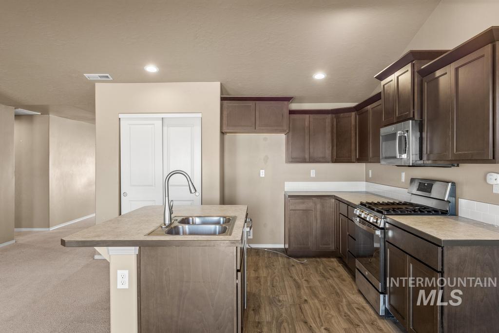 Kitchen with appliances with stainless steel finishes, a kitchen island with sink, light countertops, dark brown cabinets, and dark wood-style floors
