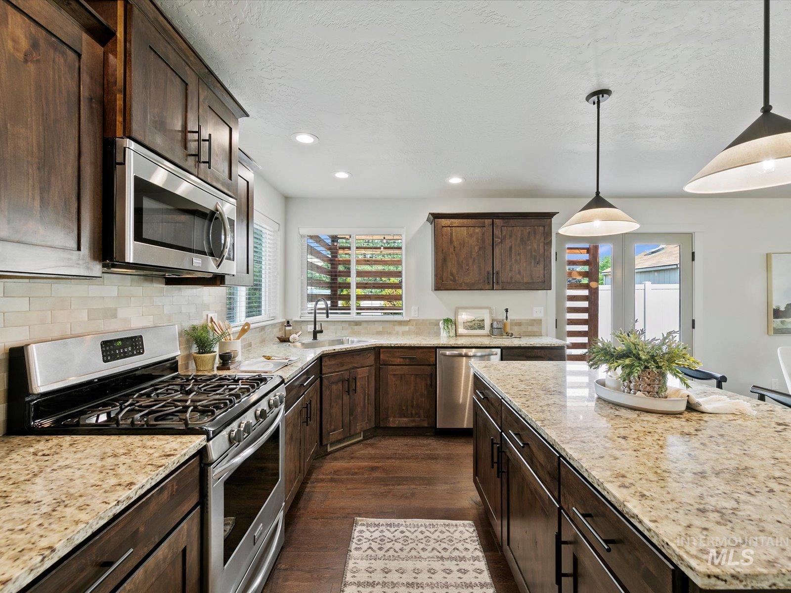 Kitchen with stainless steel appliances, dark brown cabinetry, decorative light fixtures, light stone counters, and healthy amount of natural light