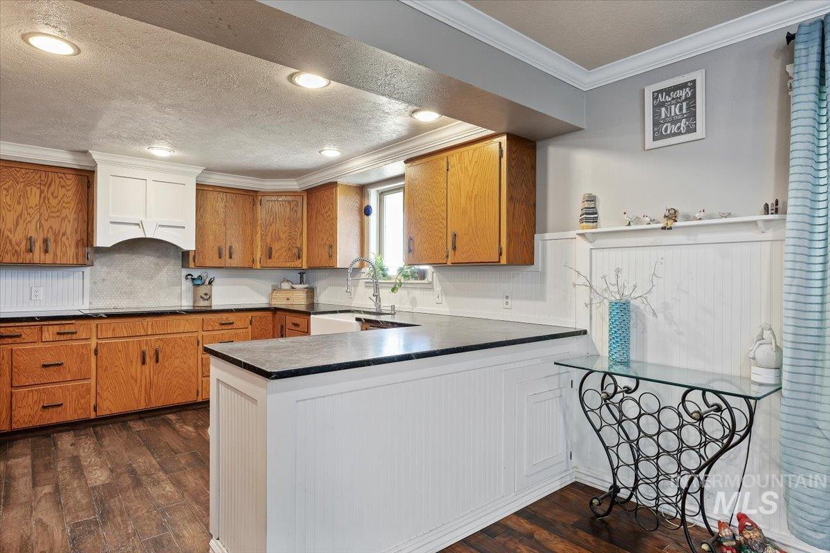 Kitchen featuring decorative backsplash, brown cabinets, ornamental molding, dark wood-style floors, and dark countertops