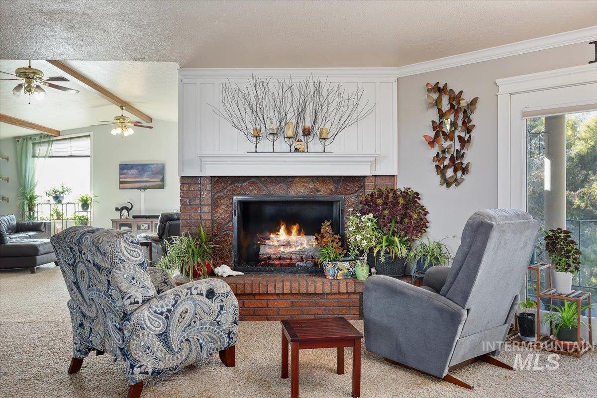 Living area featuring a textured ceiling, a brick fireplace, carpet floors, beamed ceiling, and ceiling fan