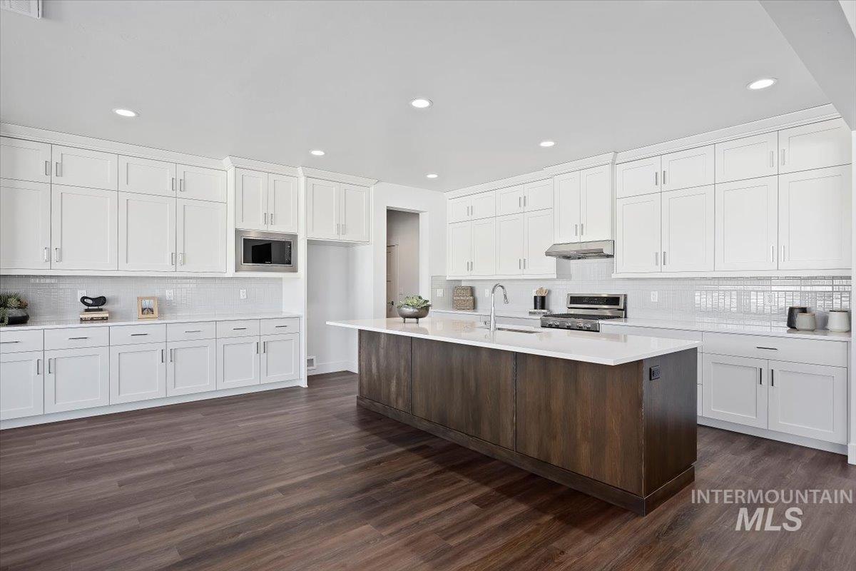 Kitchen featuring decorative backsplash, white cabinets, appliances with stainless steel finishes, a kitchen island with sink, and dark wood finished floors