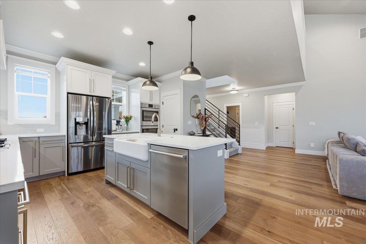 Kitchen with gray cabinetry, appliances with stainless steel finishes, an island with sink, hanging light fixtures, and light wood-style floors