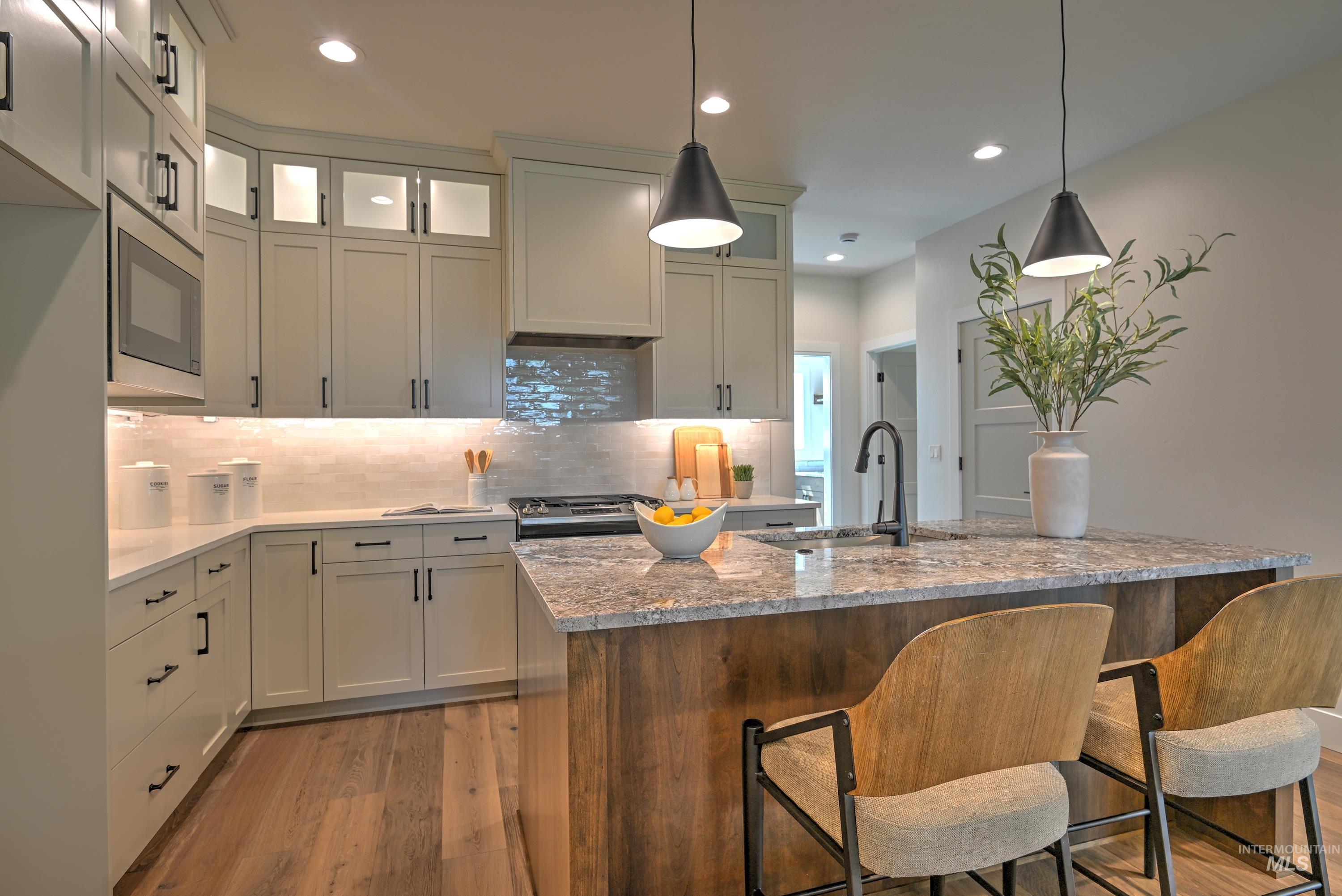 Kitchen with light quartz & granite counters, hanging light fixtures, full tile backsplash, light wood-style floors, and an island with sink