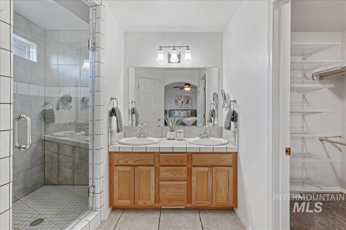 Ensuite bathroom with a walk in closet, light tile patterned flooring, double vanity, ceiling fan, and a stall shower