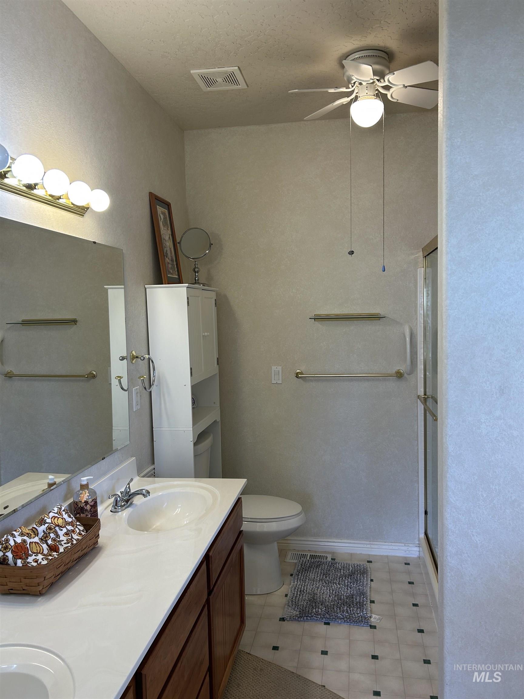 Full bath with double vanity, a shower stall, a ceiling fan, a textured wall, and a textured ceiling