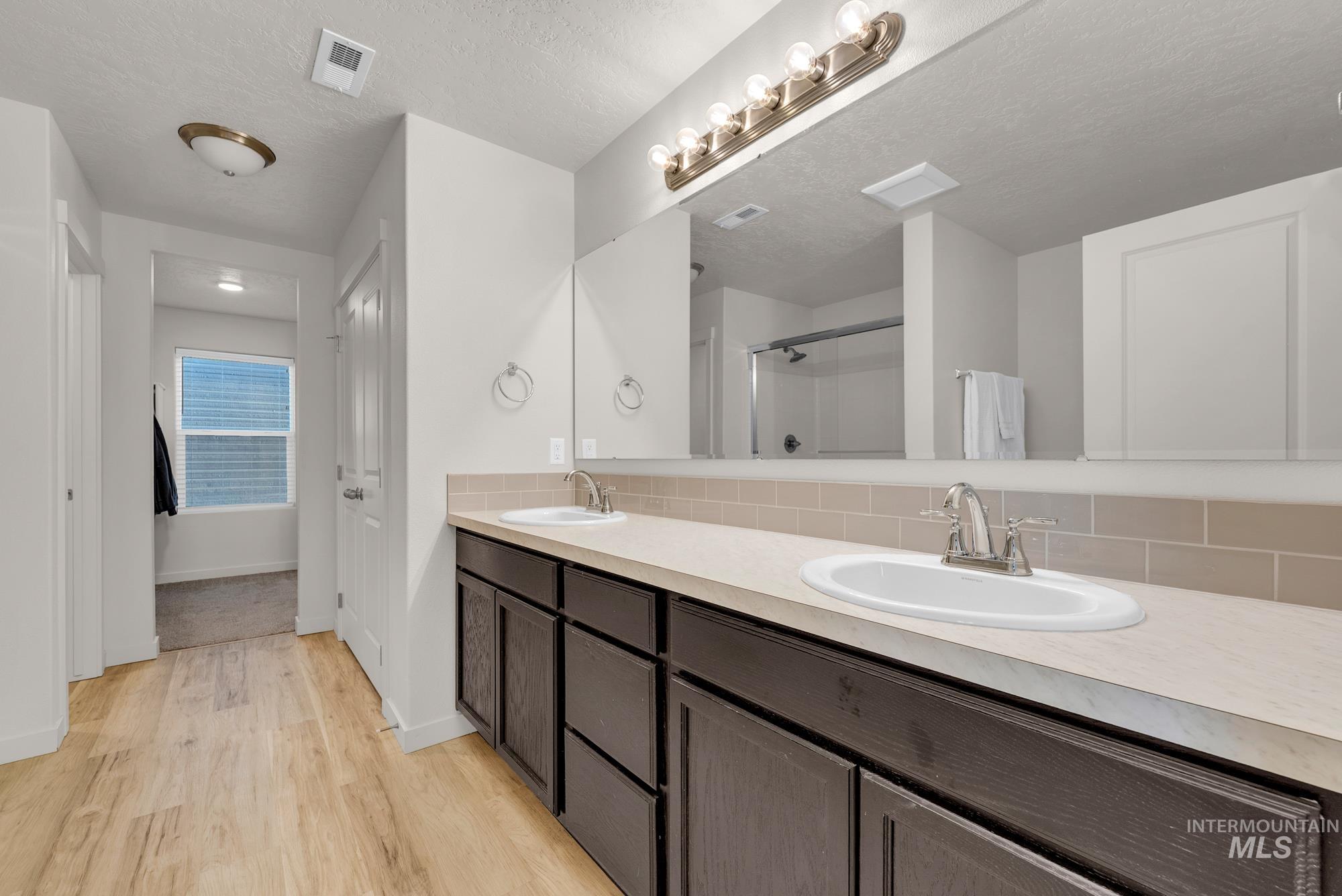 Bathroom featuring double vanity, light wood-style flooring, tasteful backsplash, a shower stall, and a textured ceiling