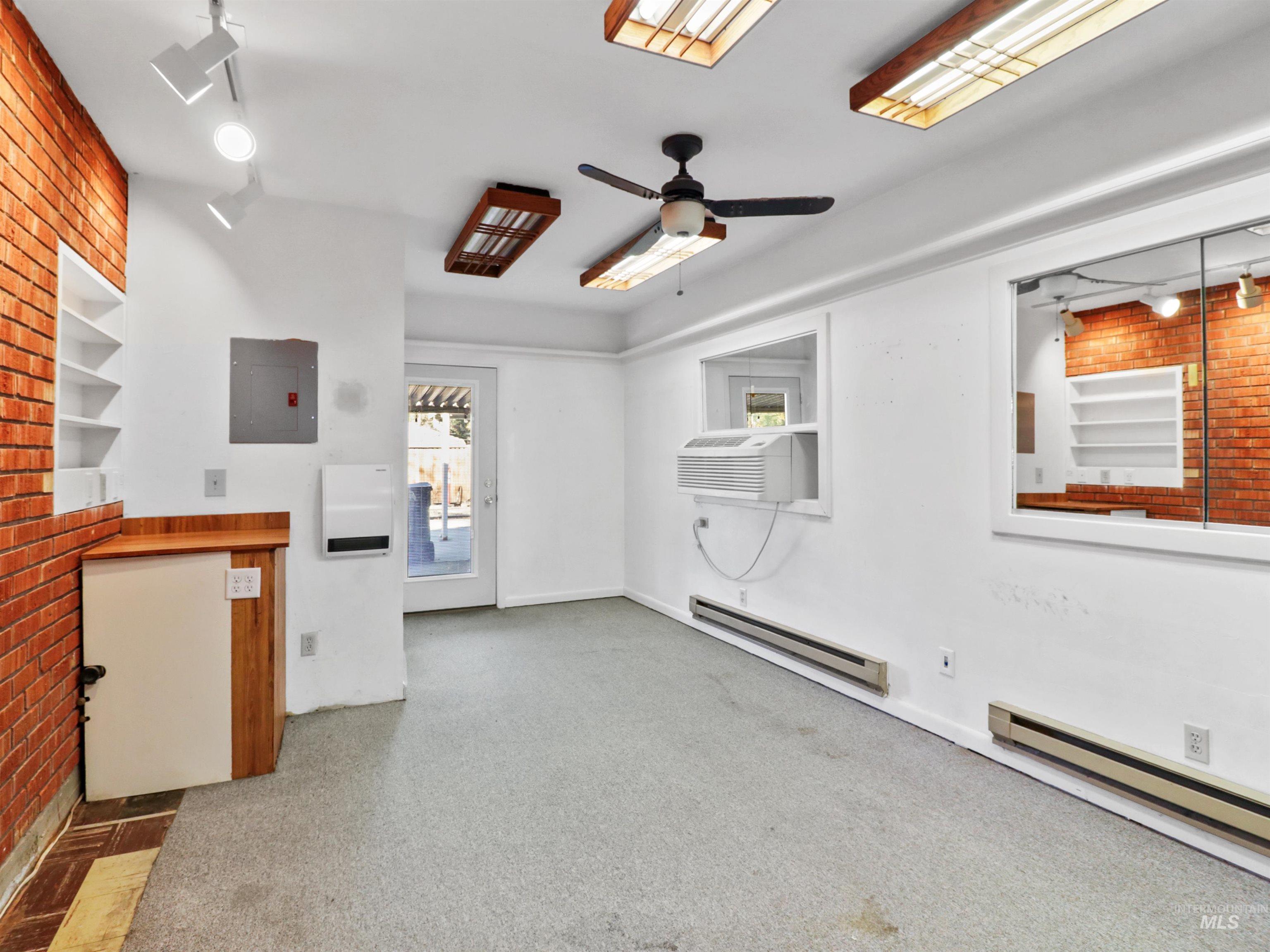 Carpeted spare room featuring electric panel, a baseboard radiator, a ceiling fan, rail lighting, and brick wall