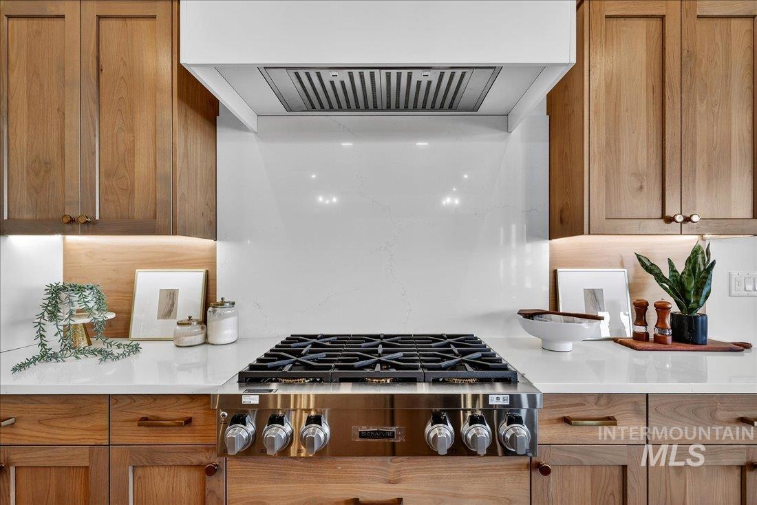 Kitchen with ventilation hood, stainless steel gas cooktop, light stone counters, and brown cabinetry