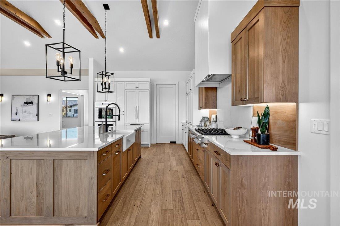 Kitchen with brown cabinetry, light wood-style floors, a large island with sink, decorative light fixtures, and light stone countertops