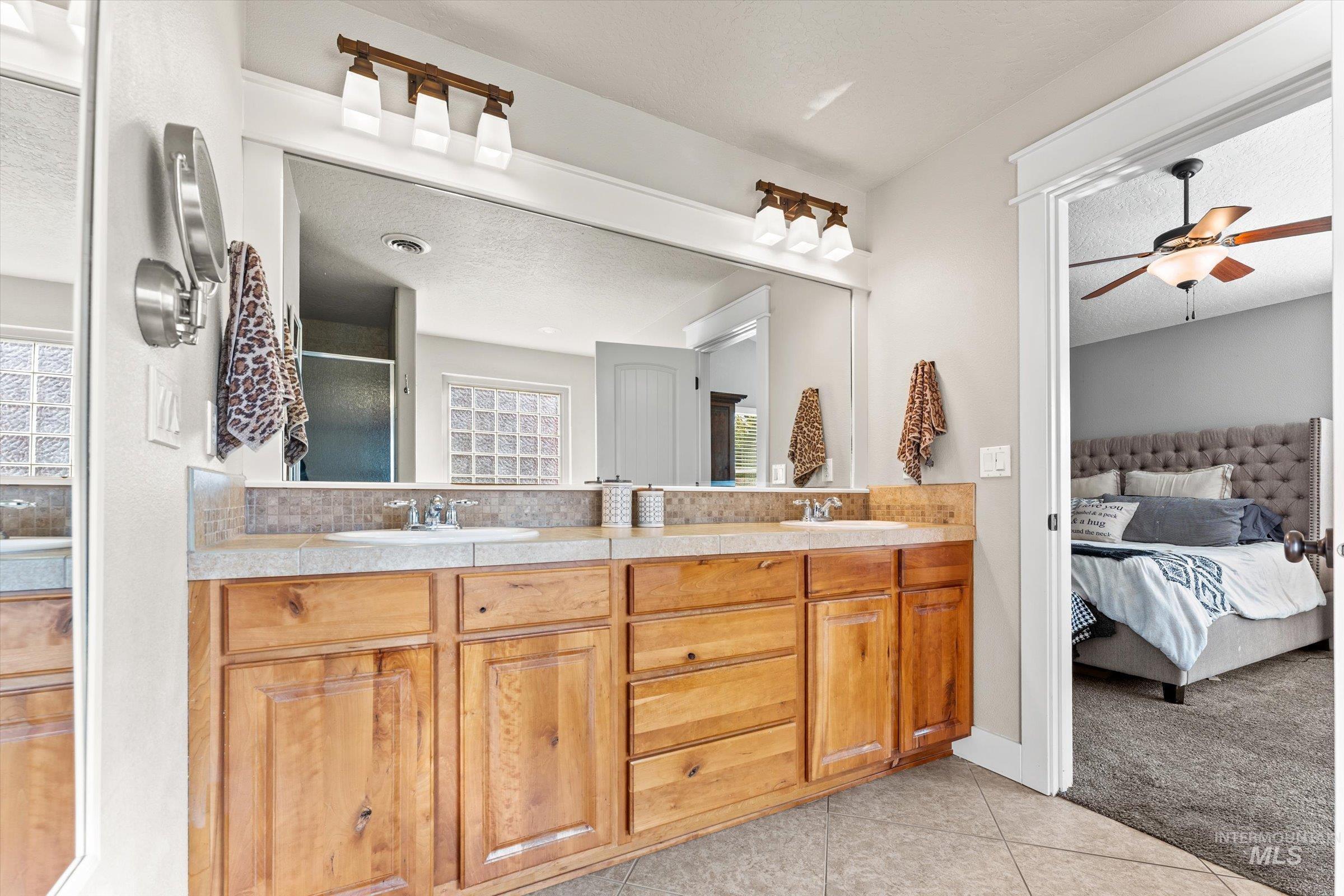 Ensuite bathroom featuring double vanity, ceiling fan, light tile patterned floors, light carpet, and a stall shower