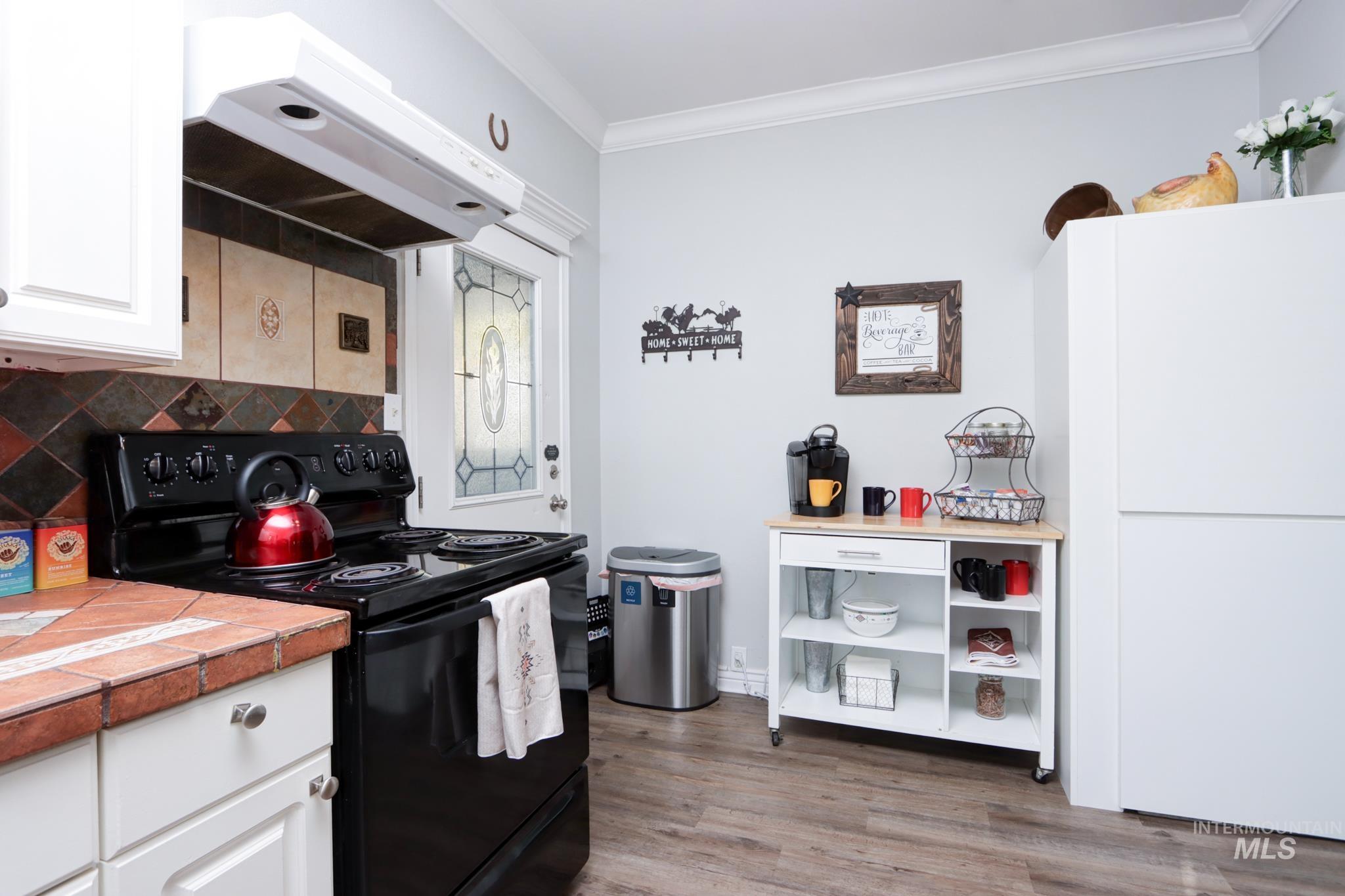 Kitchen with black electric range oven, range hood, white cabinets, ornamental molding, and light wood-style floors