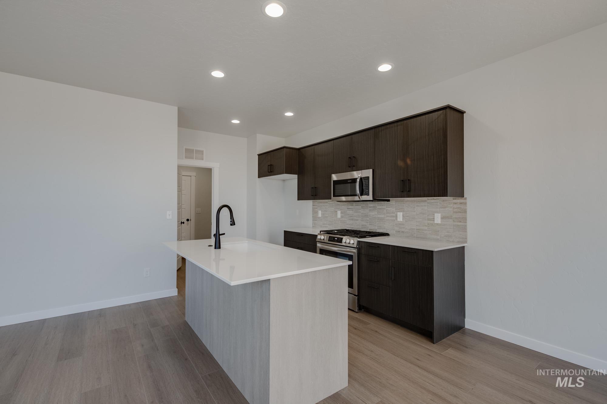 Kitchen featuring stainless steel appliances, an island with sink, backsplash, light wood-type flooring, and dark brown cabinets