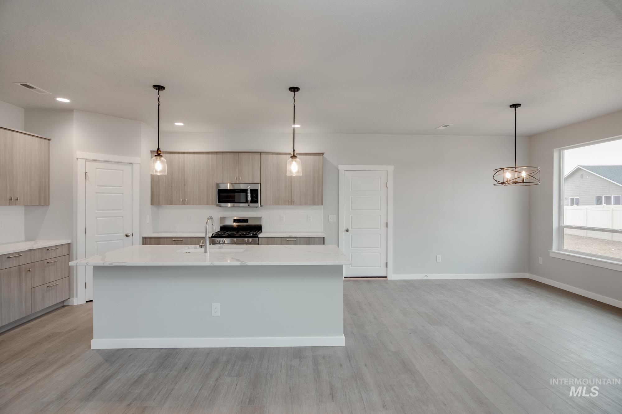 Kitchen featuring decorative light fixtures, light stone counters, stainless steel appliances, an island with sink, and light brown cabinetry