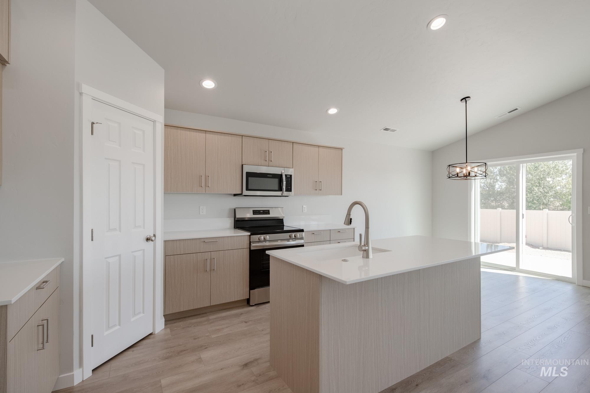 Kitchen featuring light brown cabinetry, appliances with stainless steel finishes, pendant lighting, a kitchen island with sink, and lofted ceiling