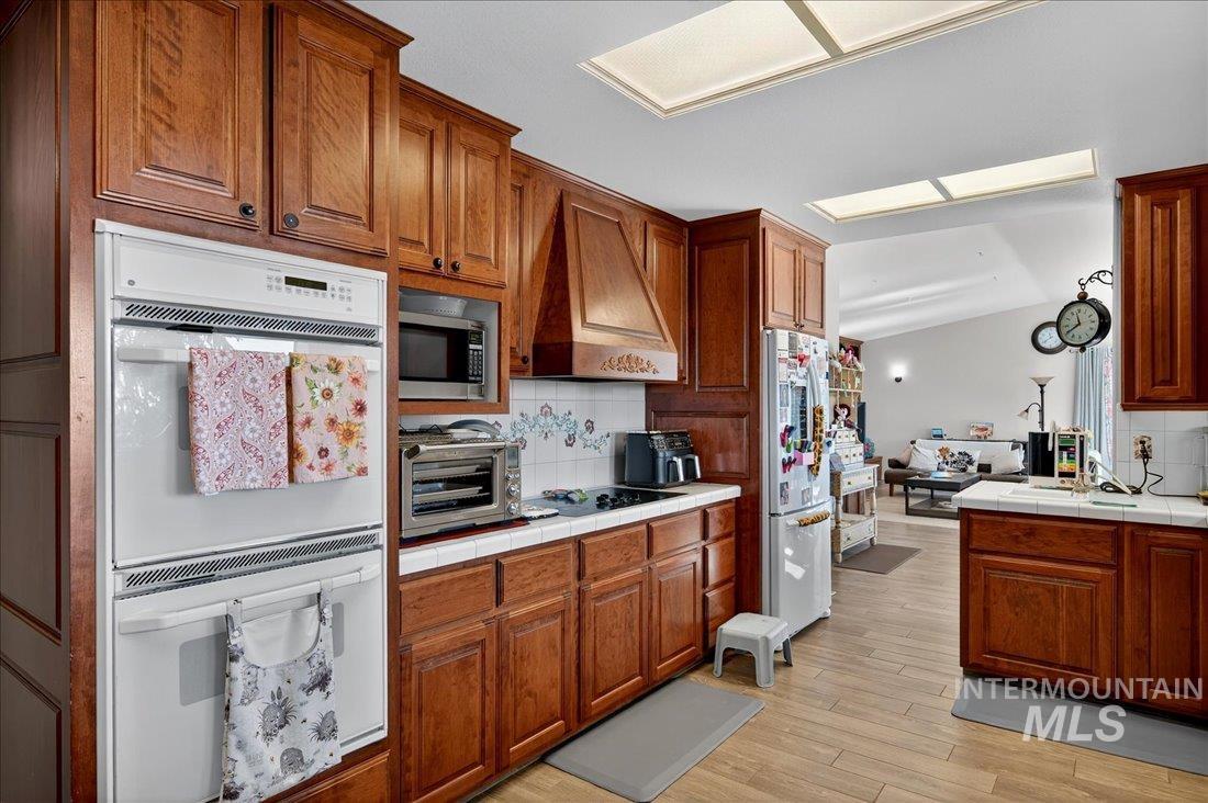 Kitchen with white appliances, backsplash, tile countertops, brown cabinetry, and light wood-style flooring