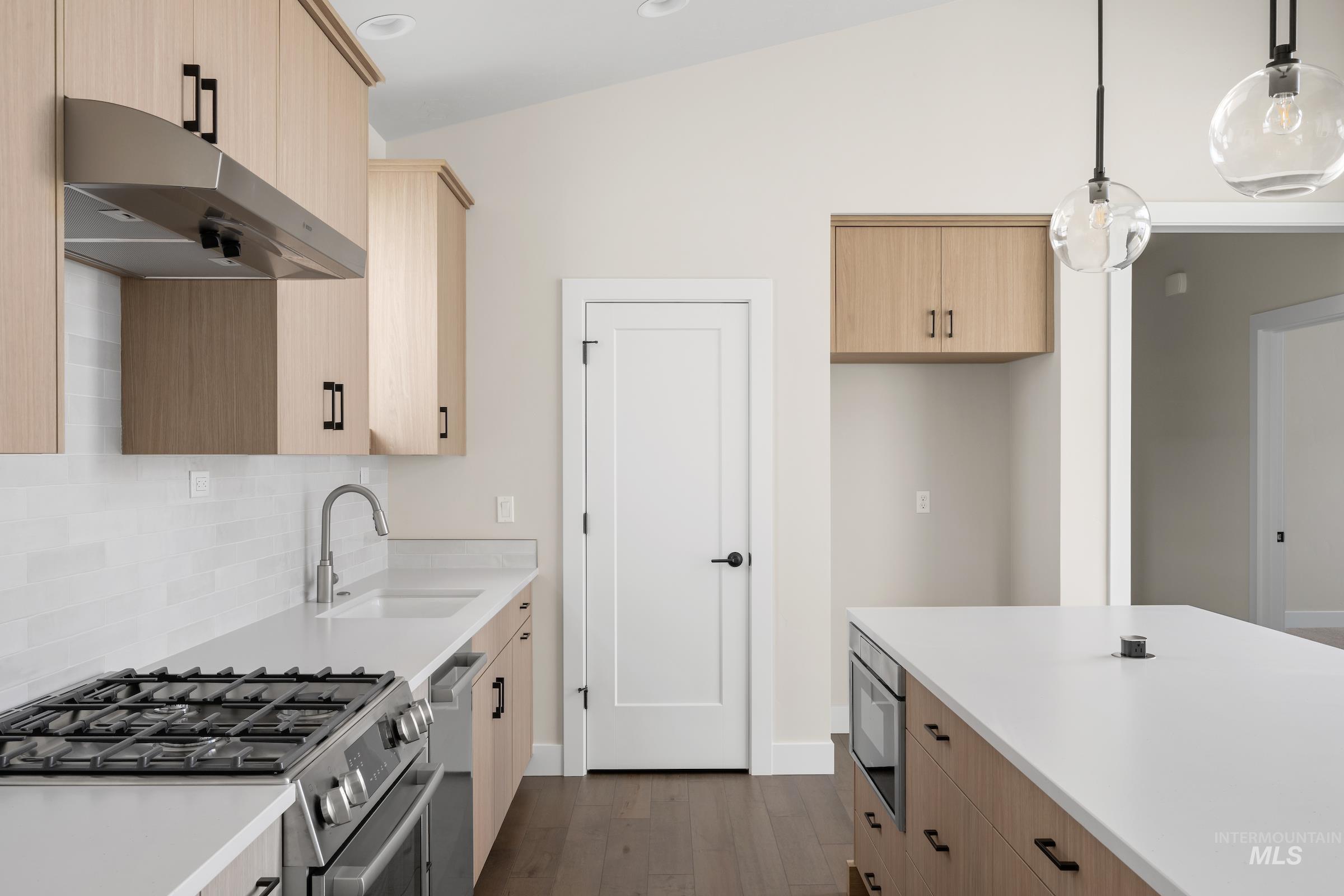 Kitchen with light brown cabinetry, stainless steel appliances, decorative light fixtures, under cabinet range hood, and dark wood-style flooring