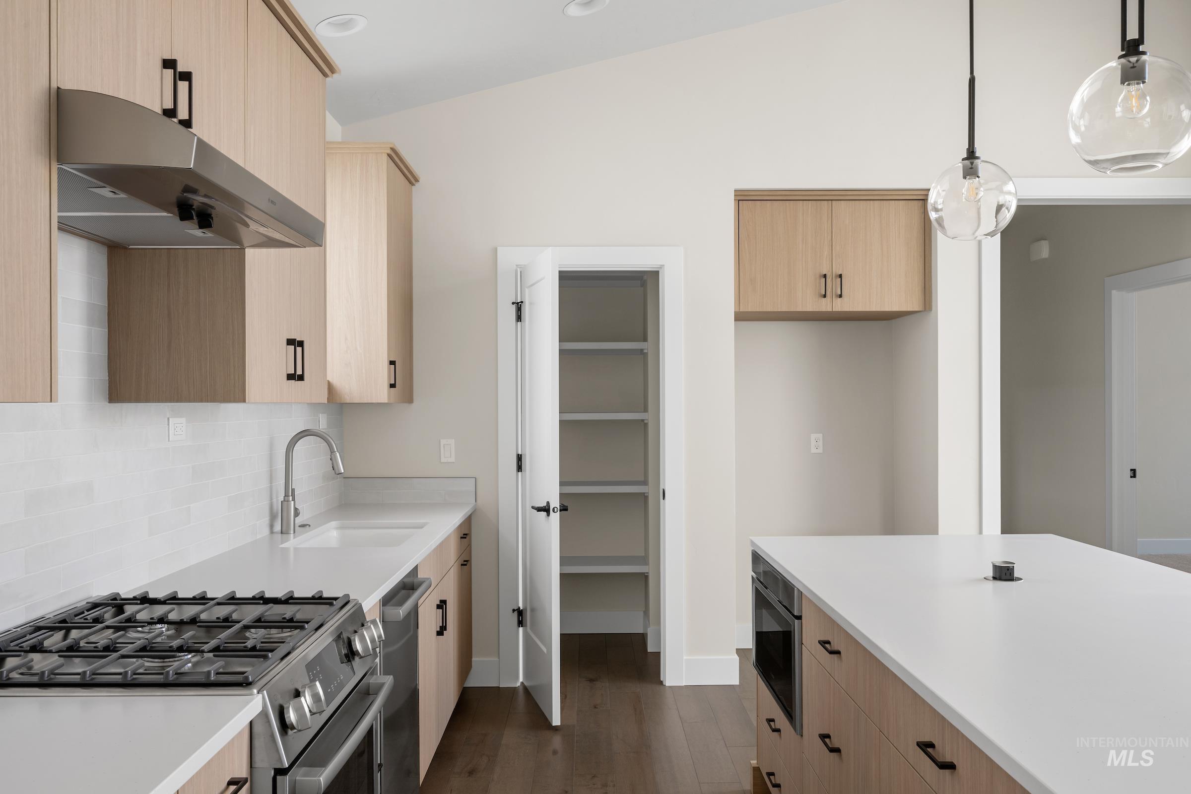 Kitchen with light brown cabinets, stainless steel appliances, pendant lighting, under cabinet range hood, and dark wood-style flooring