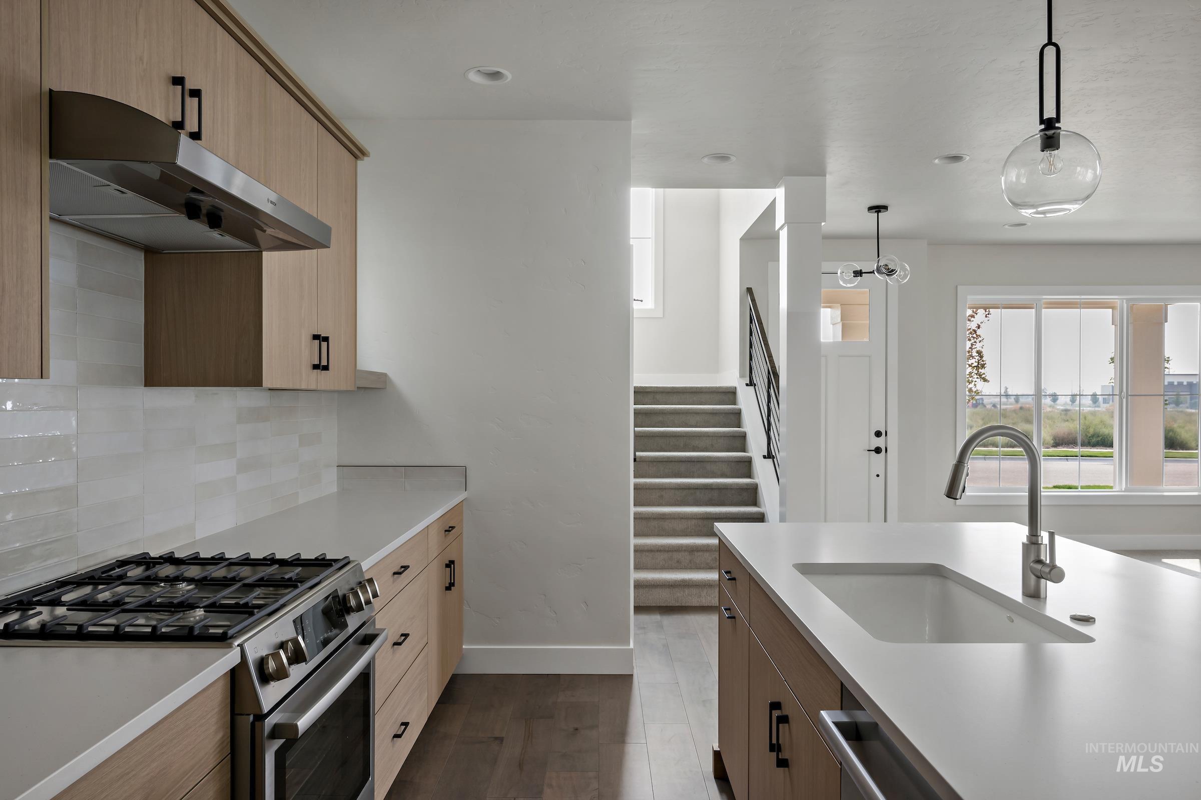 Kitchen with stainless steel appliances, under cabinet range hood, dark wood-type flooring, hanging light fixtures, and tasteful backsplash
