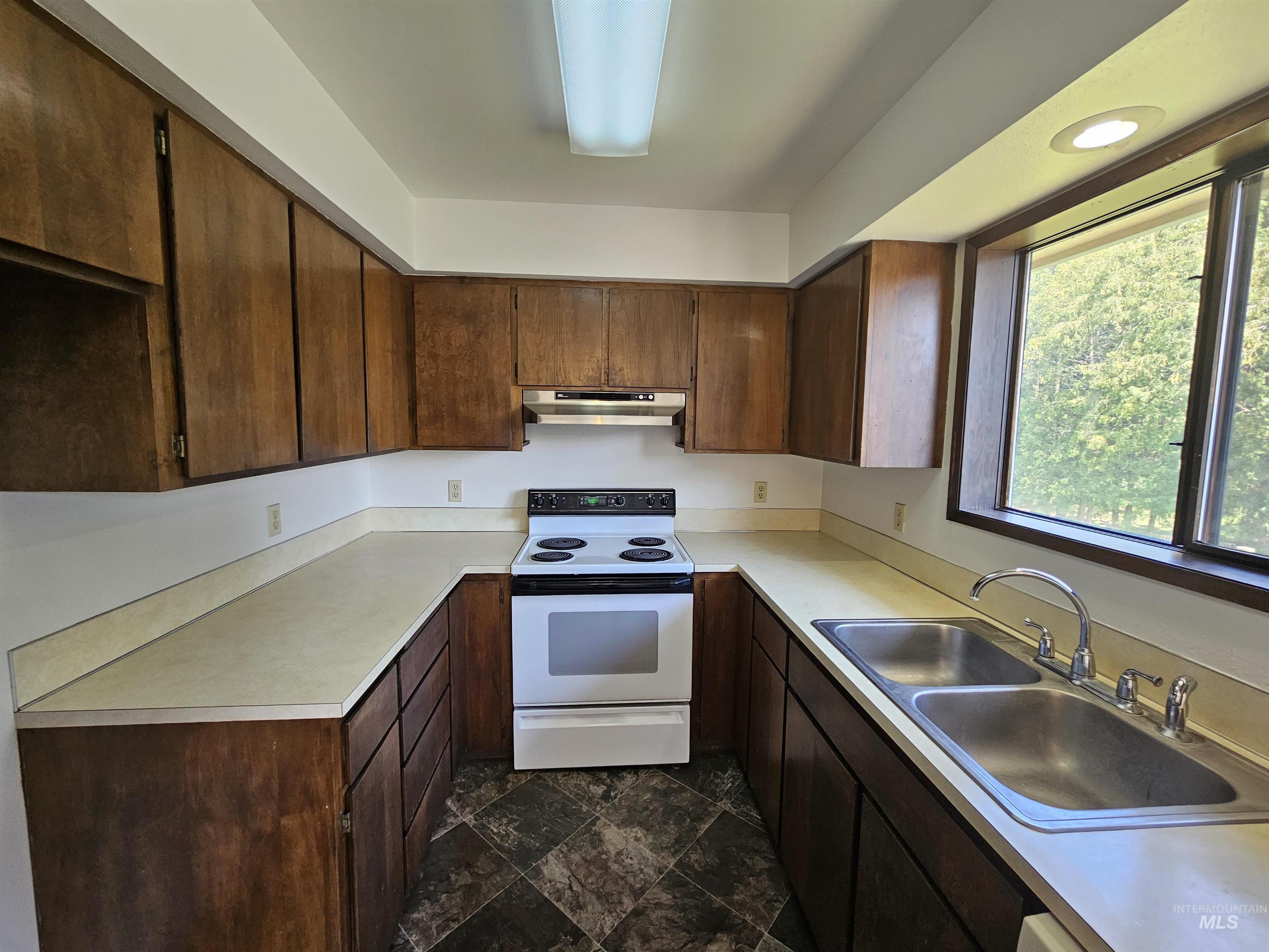 Kitchen with white electric stove, light countertops, dark brown cabinets, under cabinet range hood, and dark stone finish flooring