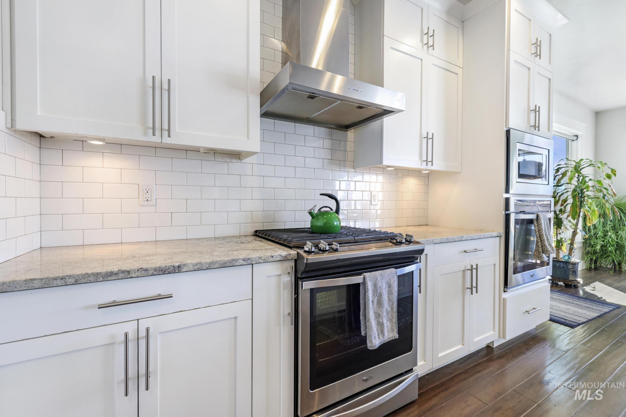 Kitchen featuring appliances with stainless steel finishes, wall chimney exhaust hood, white cabinets, light stone countertops, and dark wood-style flooring