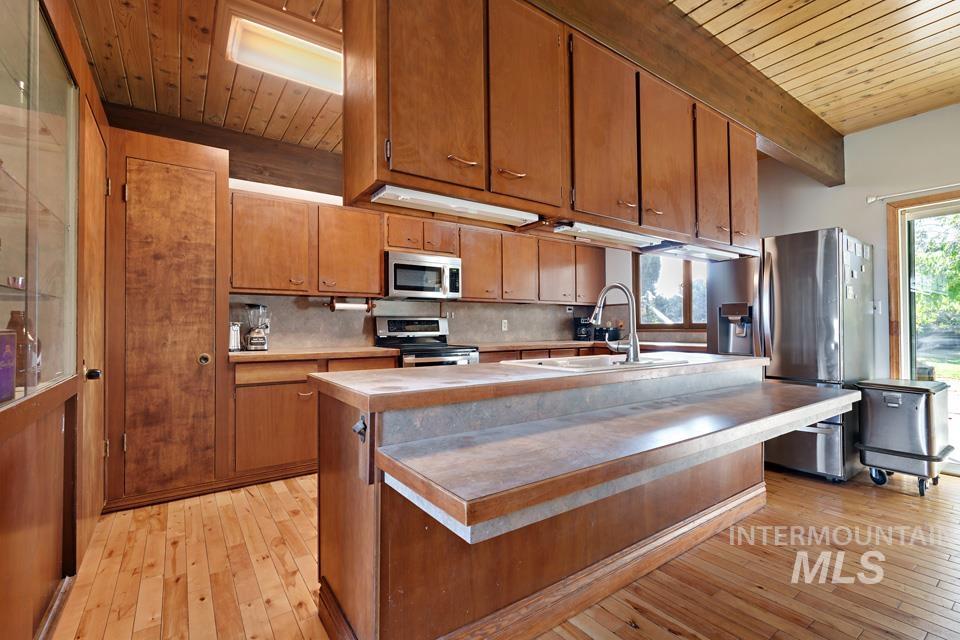 Kitchen featuring a wood ceiling with exposed beams, light countertops, appliances with stainless steel finishes, a center island with sink, and brown cabinets