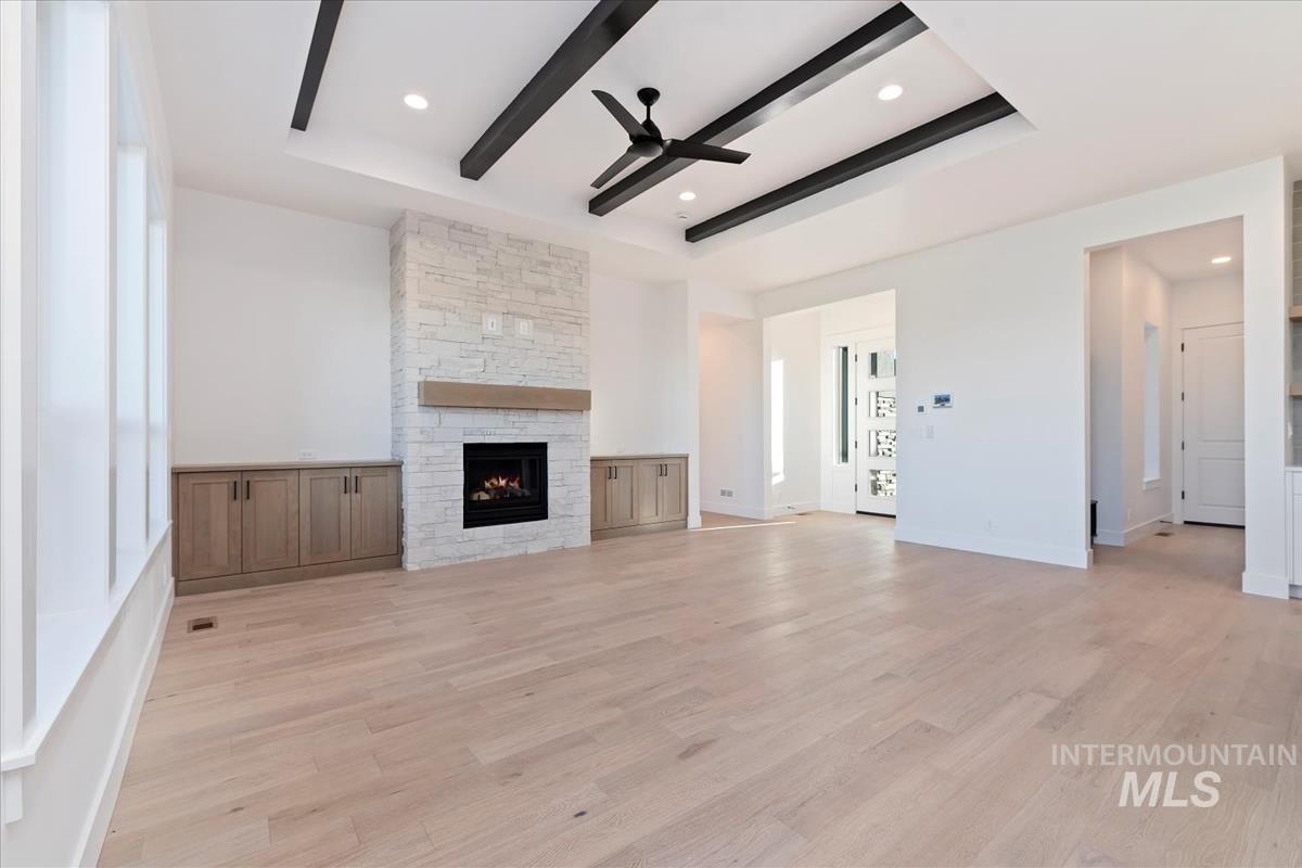 Unfurnished living room with light wood finished floors, a stone fireplace, a tray ceiling, a ceiling fan, and beamed ceiling