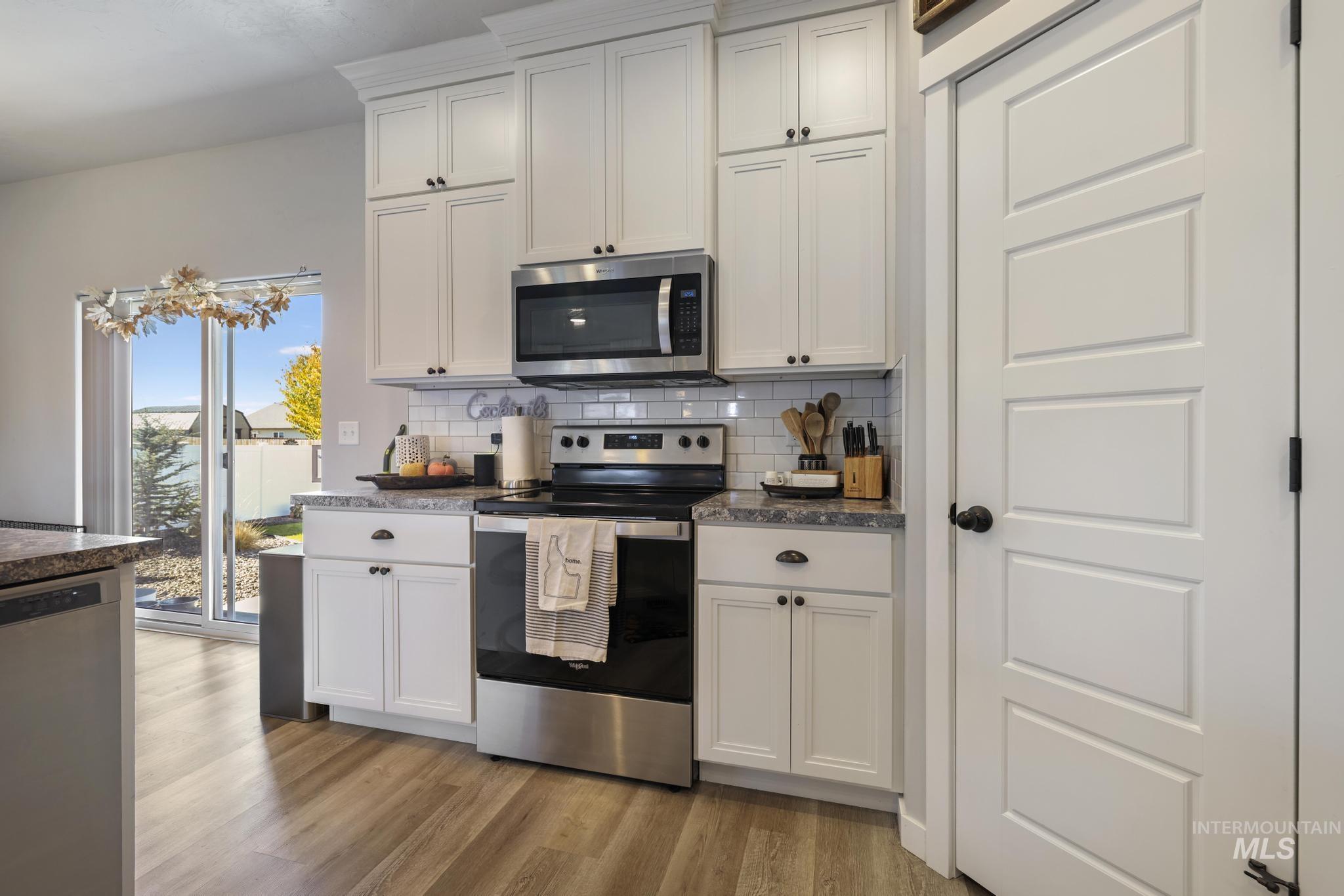 Kitchen with stainless steel appliances, tasteful backsplash, white cabinets, light wood finished floors, and dark stone counters