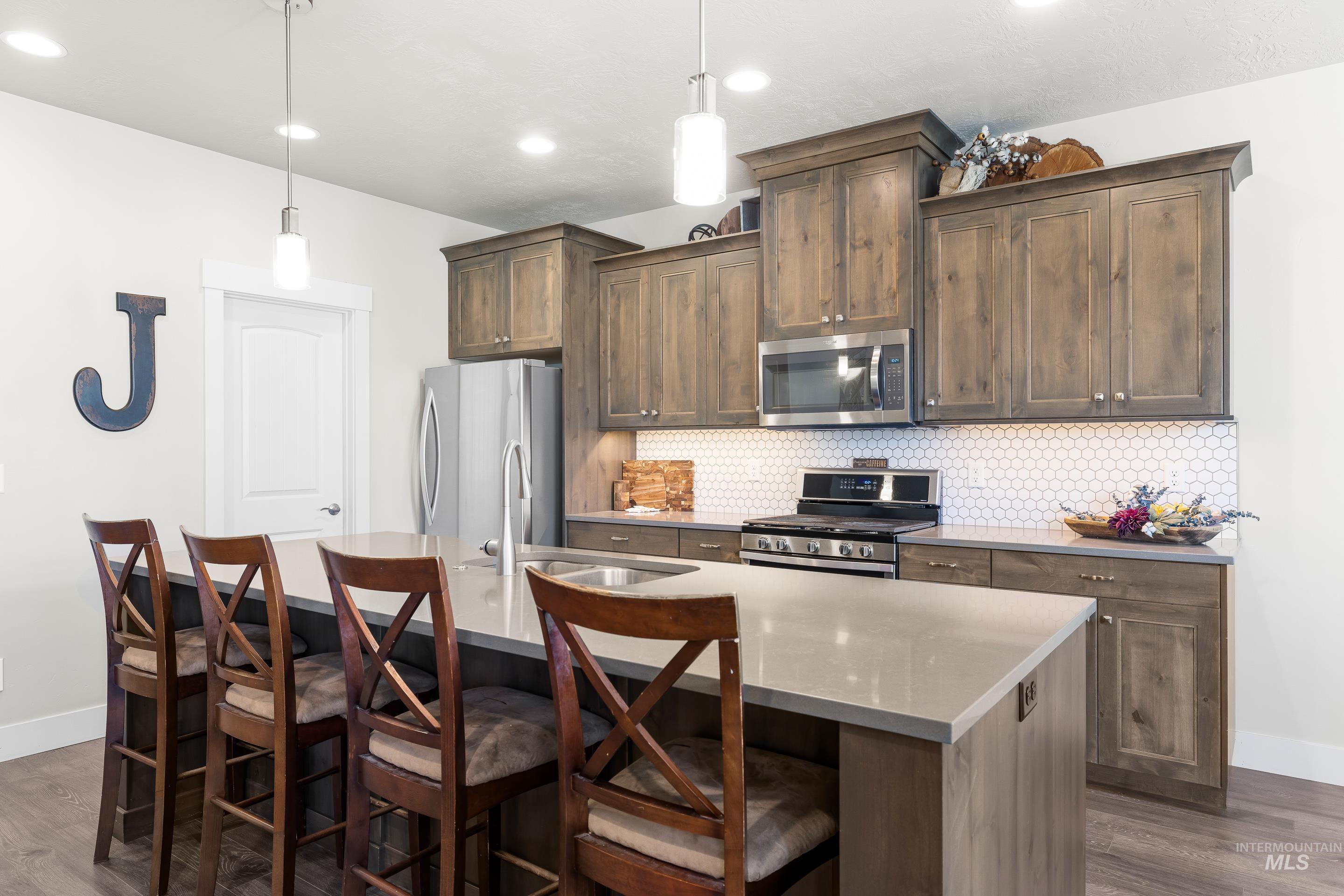 Kitchen with dark wood finished floors, backsplash, decorative light fixtures, appliances with stainless steel finishes, and an island with sink