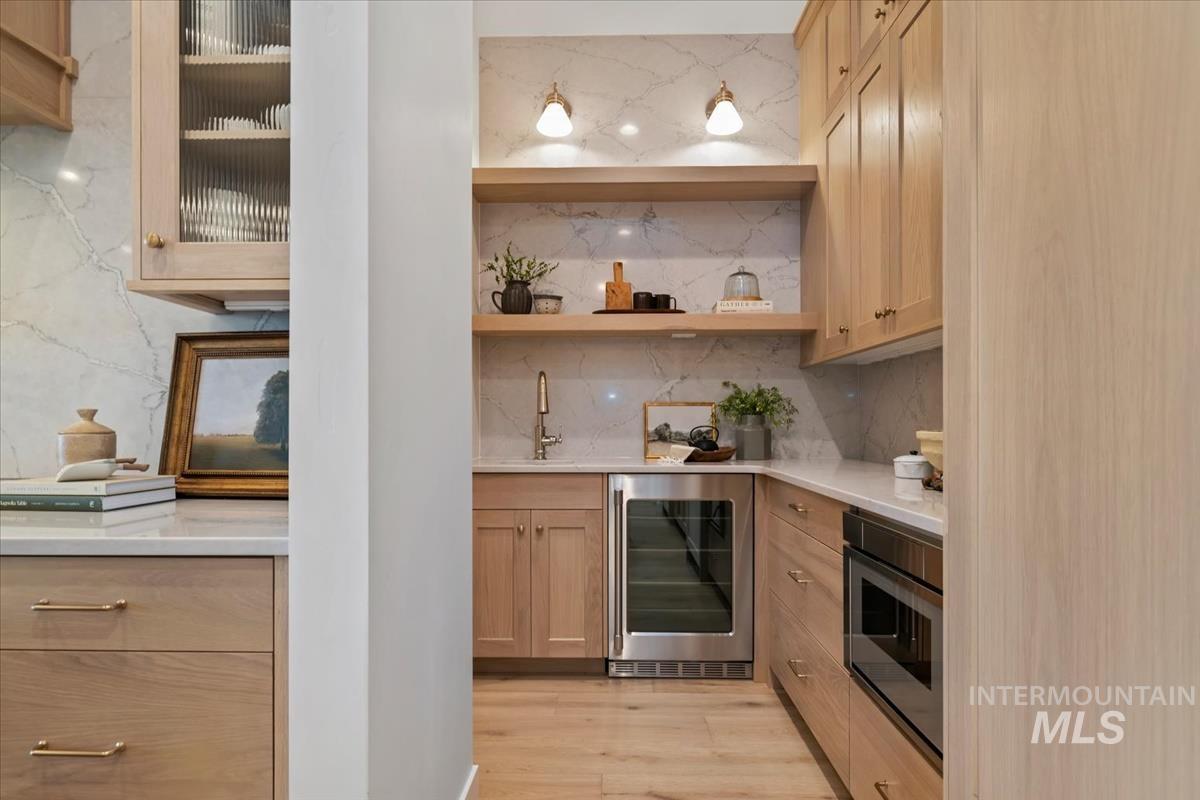 Bar area featuring wine cooler, light wood-style flooring, light stone counters, light brown cabinetry, and backsplash