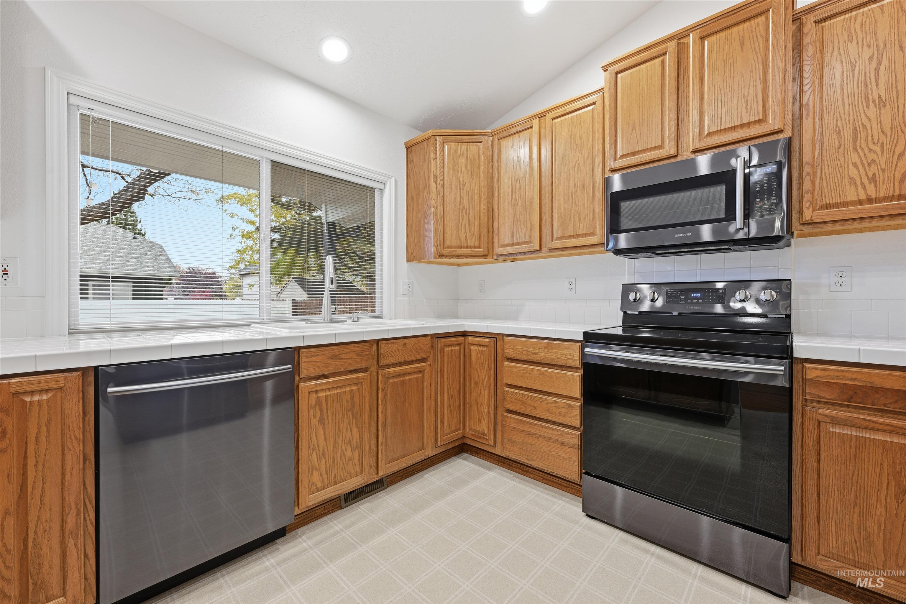 Kitchen featuring tile counters, appliances with stainless steel finishes, brown cabinets, recessed lighting, and vaulted ceiling