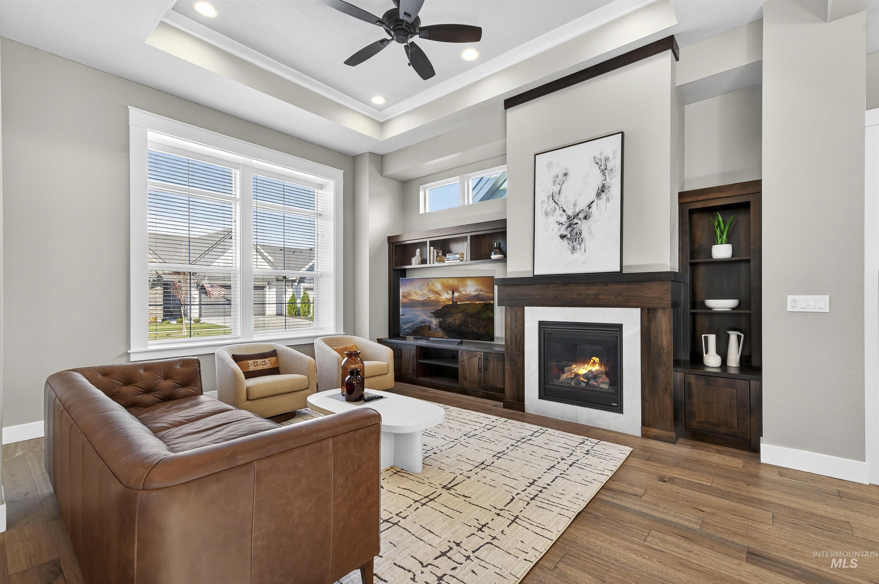 Living room featuring wood finished floors, a glass covered fireplace, a raised ceiling, a ceiling fan, and recessed lighting