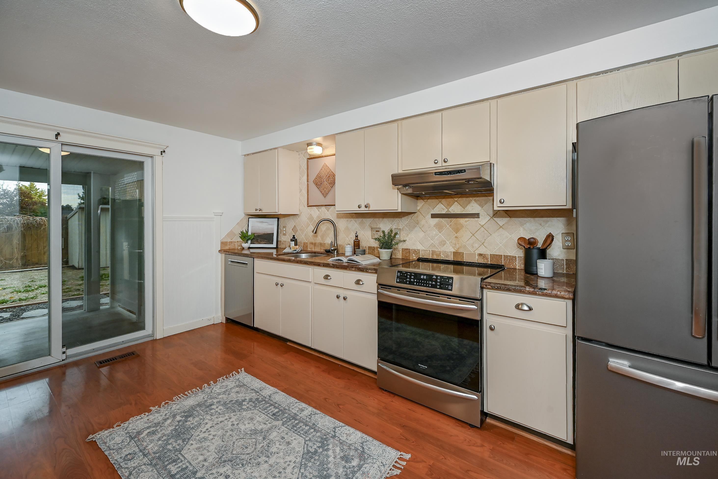 Kitchen featuring appliances with stainless steel finishes, dark wood-type flooring, cream cabinetry, decorative backsplash, and under cabinet range hood