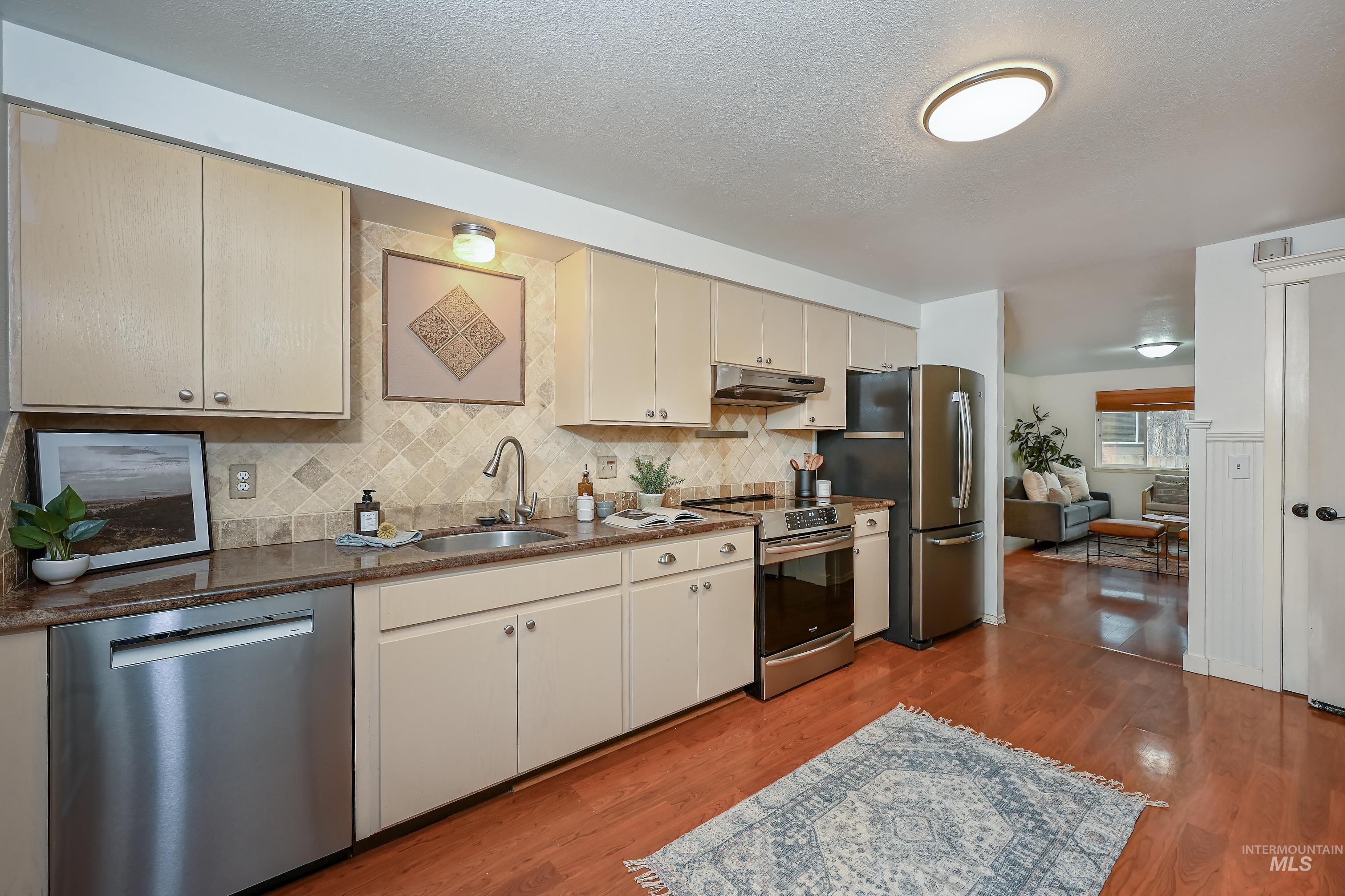 Kitchen with appliances with stainless steel finishes, tasteful backsplash, light wood finished floors, under cabinet range hood, and dark stone counters