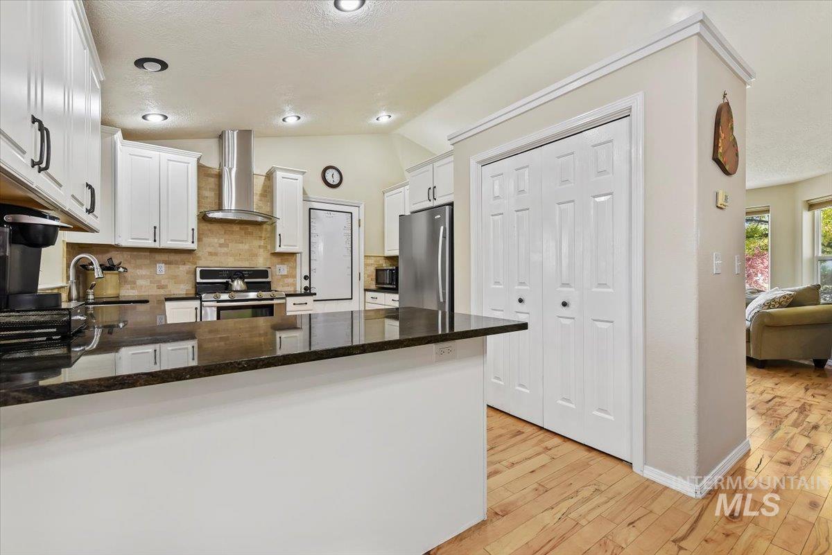 Kitchen with dark stone counters, backsplash, wall chimney exhaust hood, white cabinetry, and appliances with stainless steel finishes