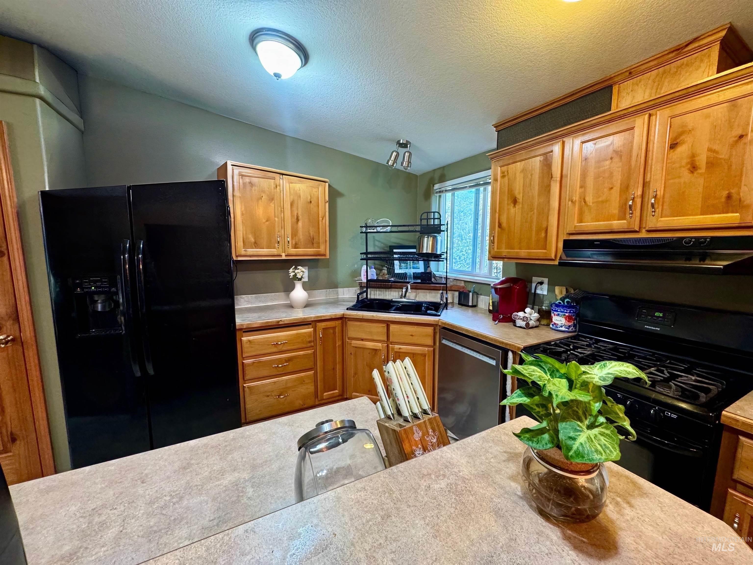 Kitchen with black appliances, light countertops, brown cabinetry, a textured ceiling, and under cabinet range hood
