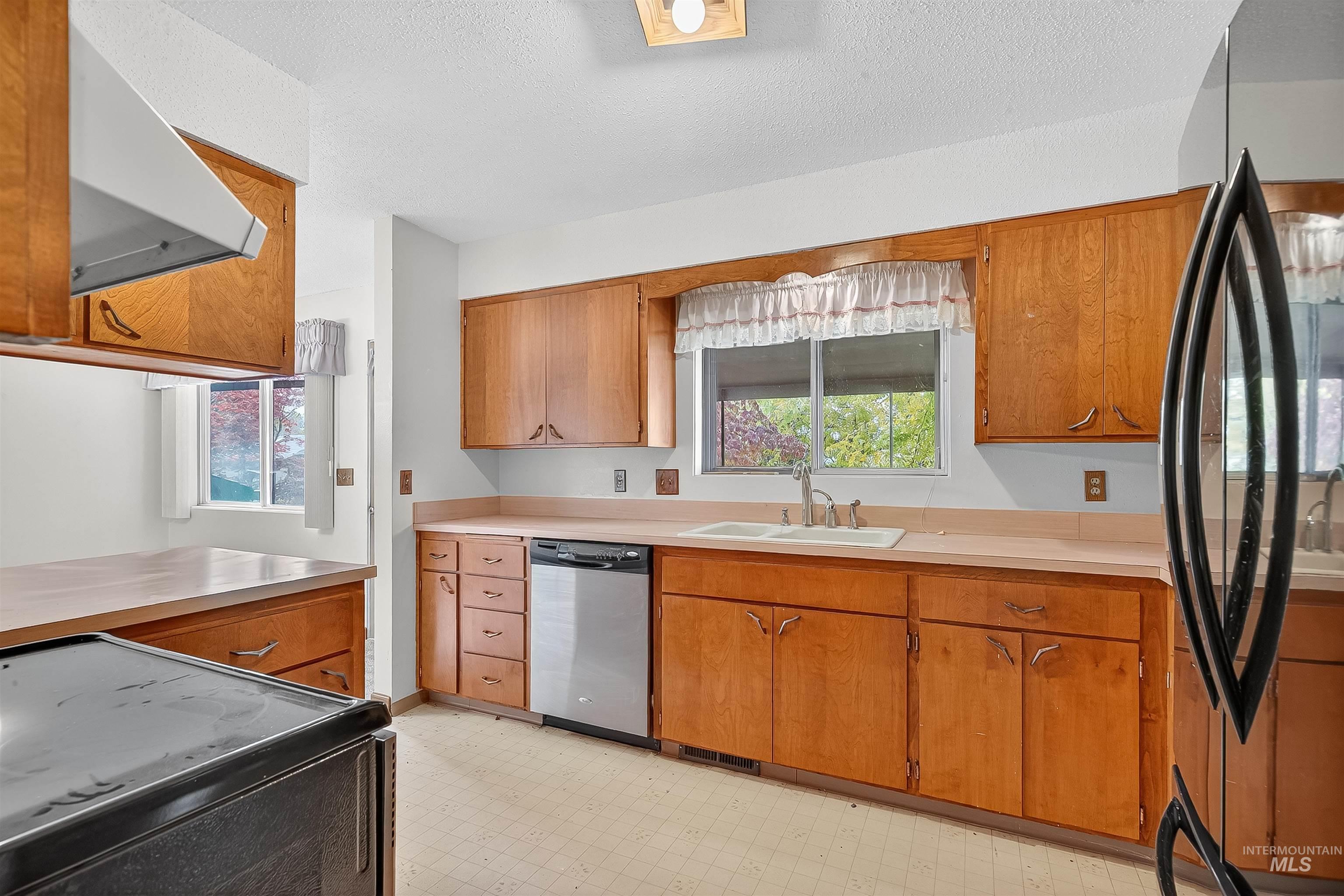 Kitchen featuring black appliances, light countertops, light floors, range hood, and brown cabinetry
