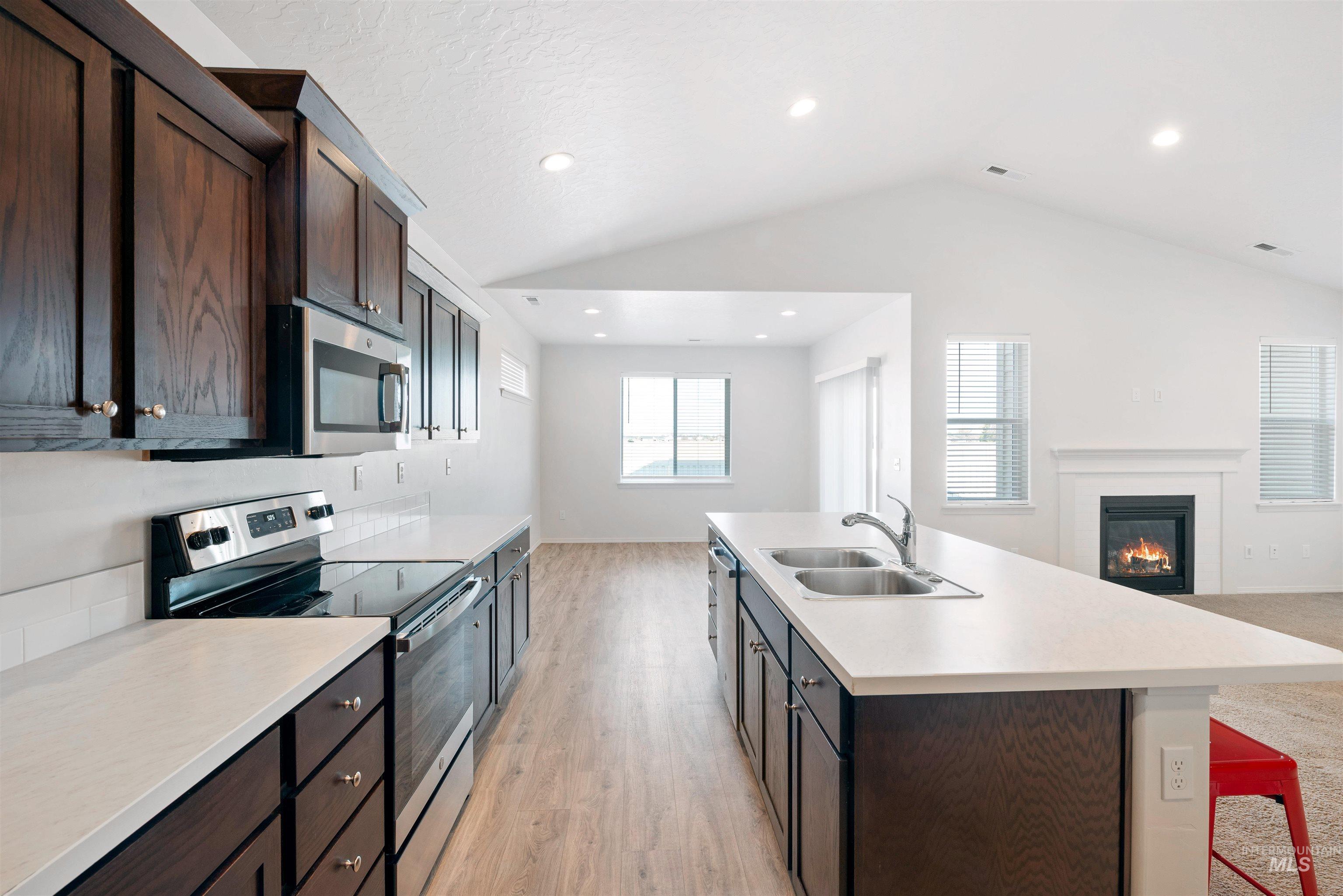 Kitchen featuring stainless steel appliances, open floor plan, dark brown cabinetry, vaulted ceiling, and a glass covered fireplace