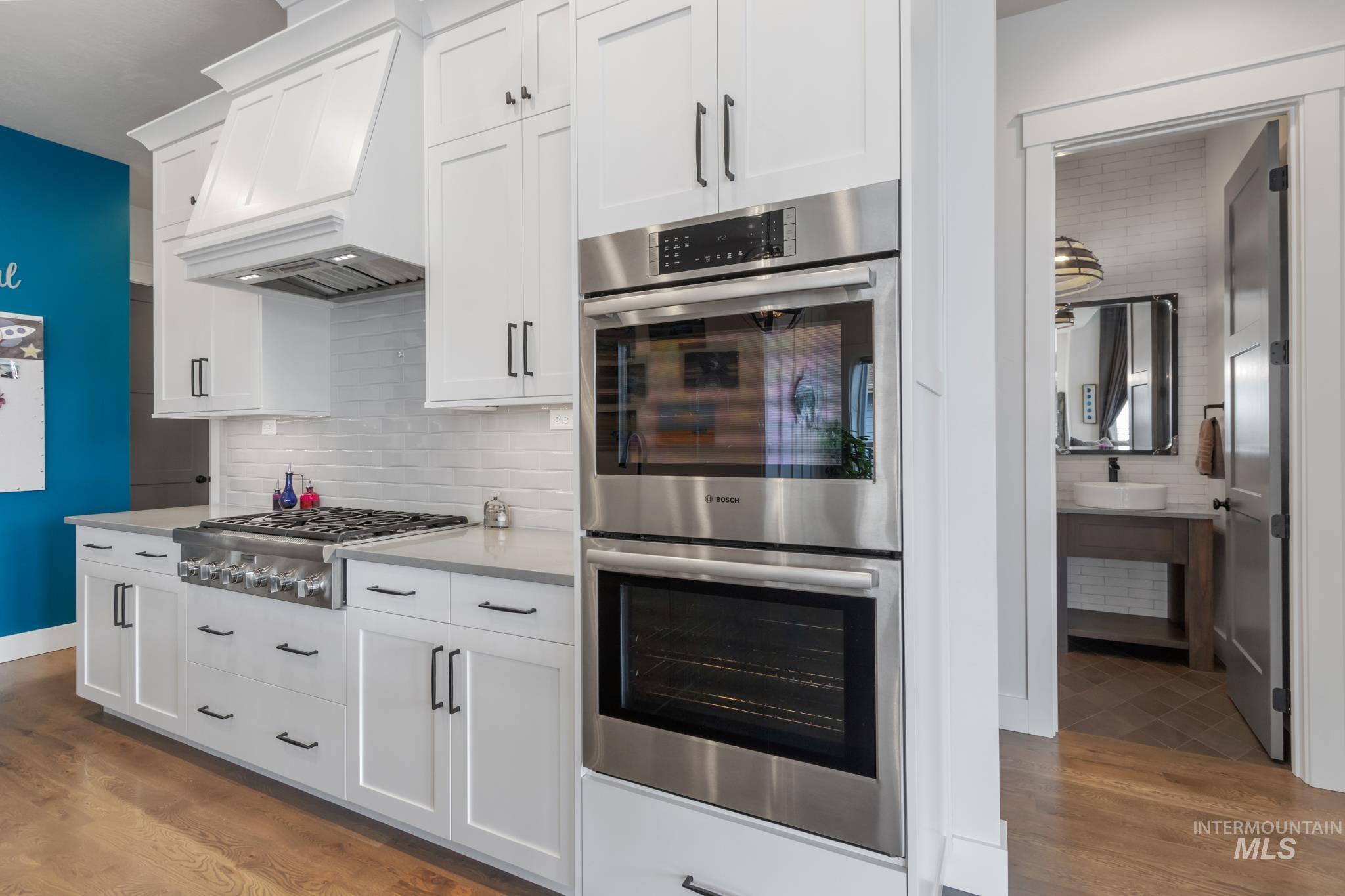 Kitchen with stainless steel appliances, dark wood-style floors, custom range hood, white cabinetry, and tasteful backsplash