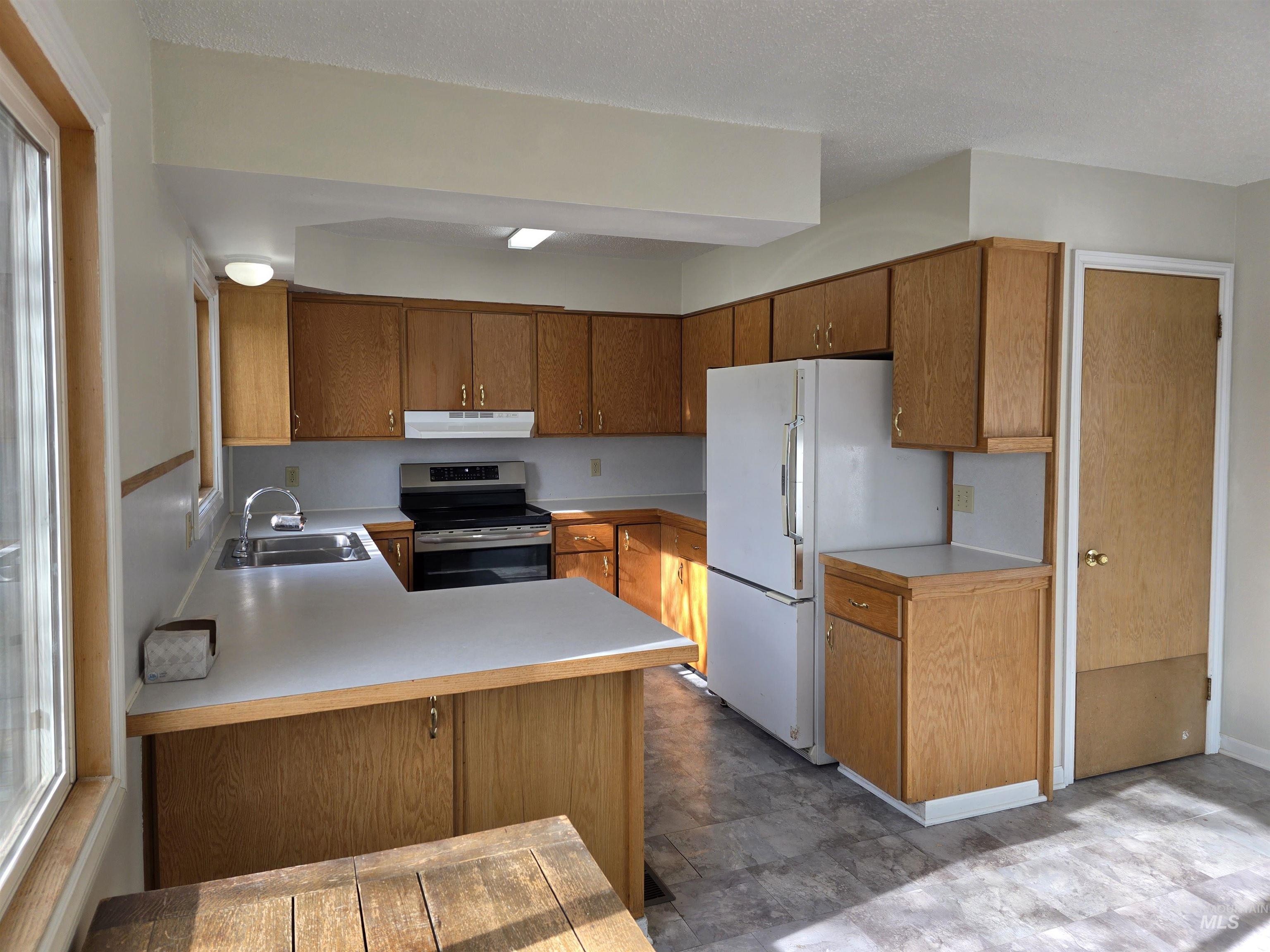 Kitchen featuring brown cabinetry, light countertops, freestanding refrigerator, stainless steel electric range oven, and a peninsula