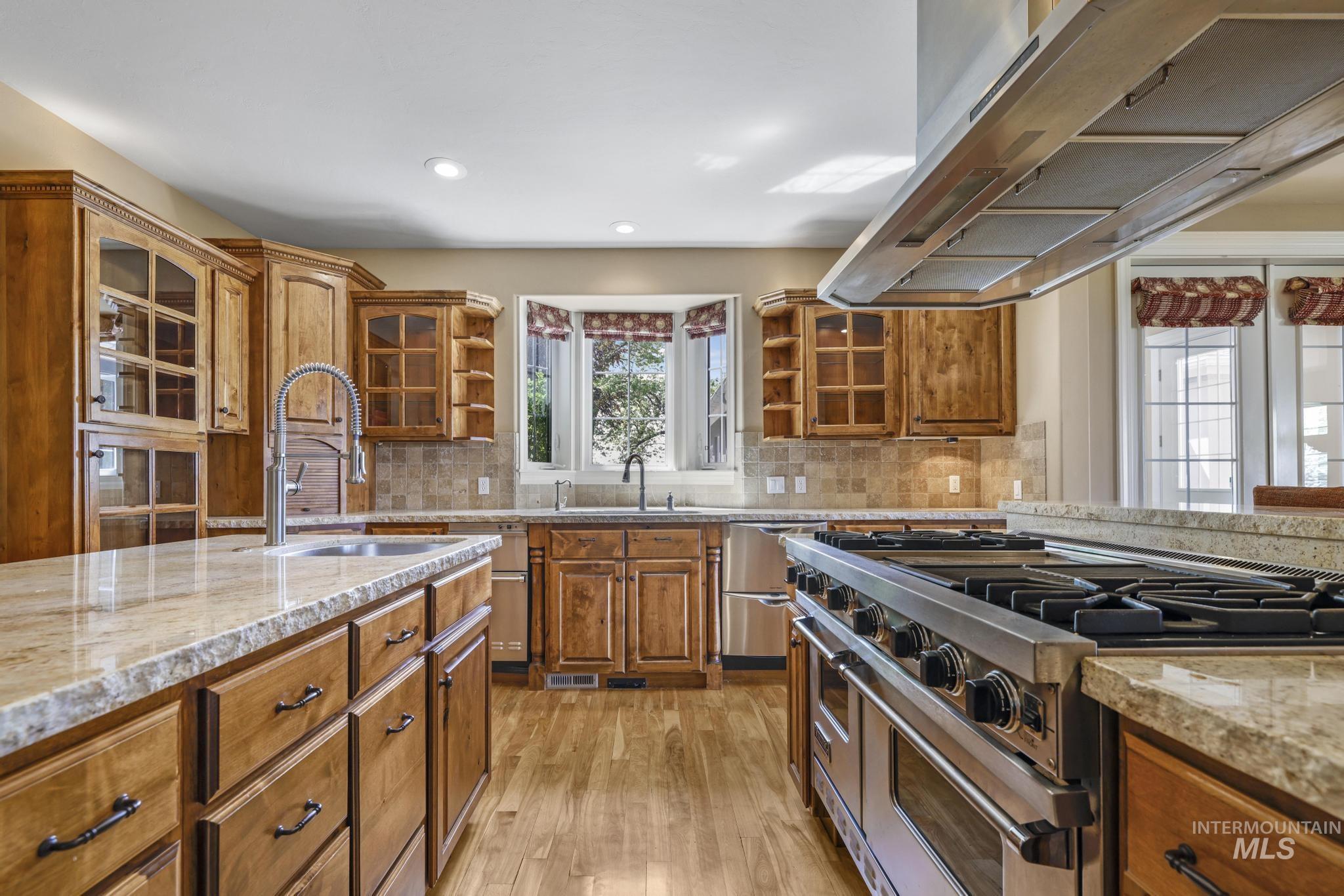 Kitchen with brown cabinetry, appliances with stainless steel finishes, light stone countertops, extractor fan, and glass insert cabinets