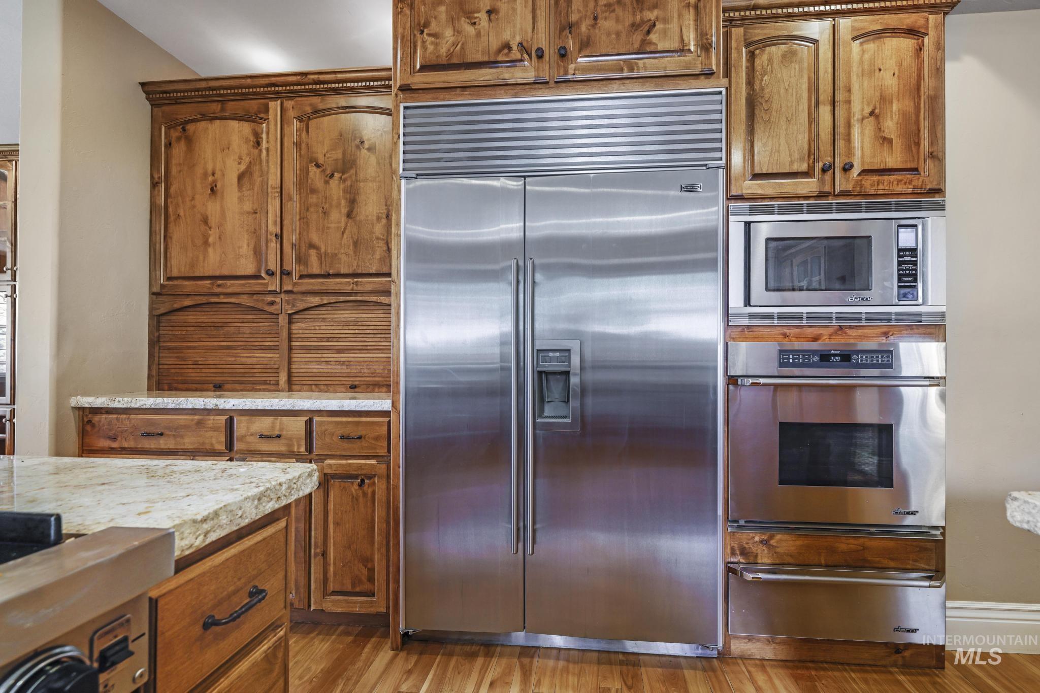 Kitchen with brown cabinets, built in appliances, a warming drawer, light wood-type flooring, and light stone countertops