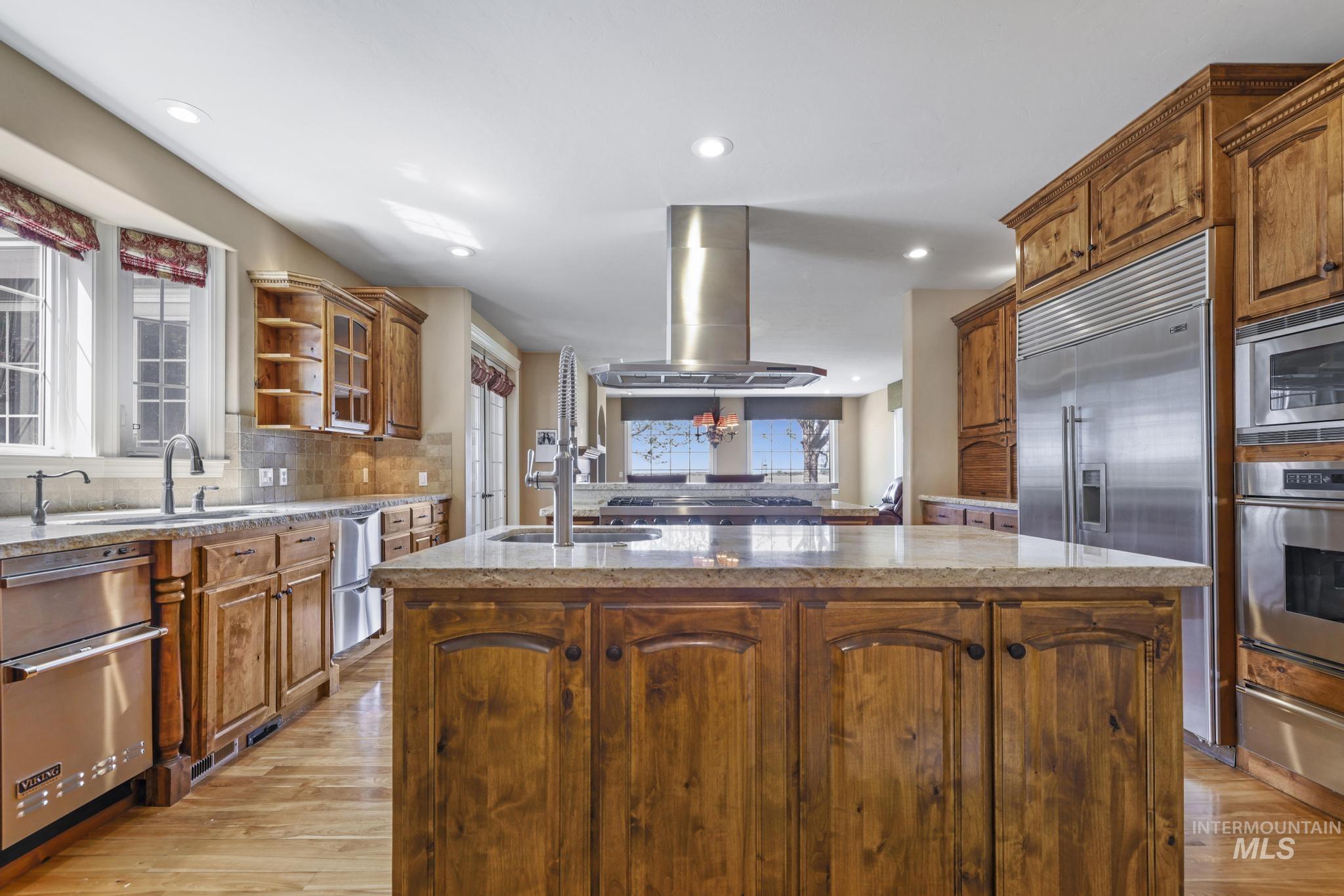Kitchen featuring a kitchen island with sink, brown cabinetry, light stone countertops, built in appliances, and light wood-style floors