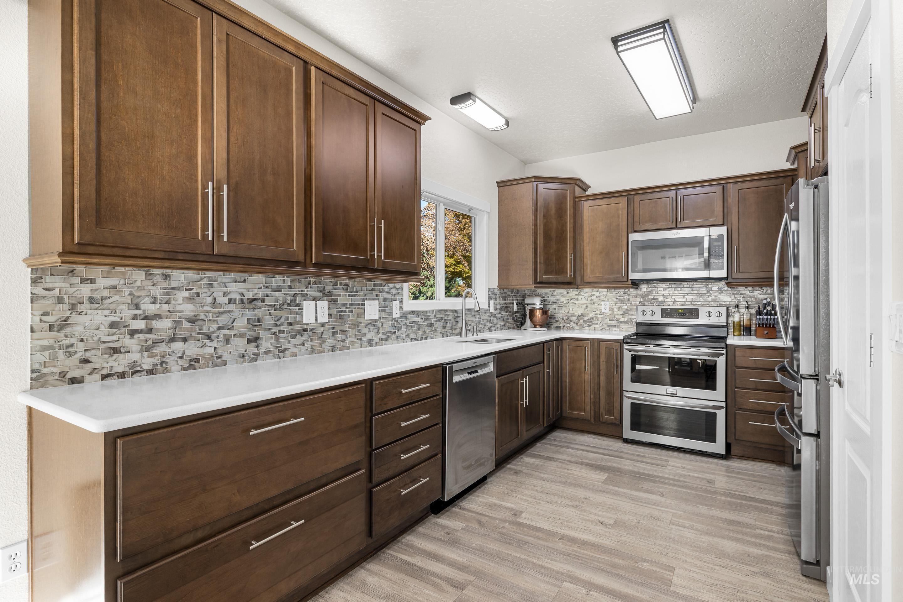 Kitchen featuring appliances with stainless steel finishes, light wood-type flooring, backsplash, dark brown cabinetry, and light stone counters