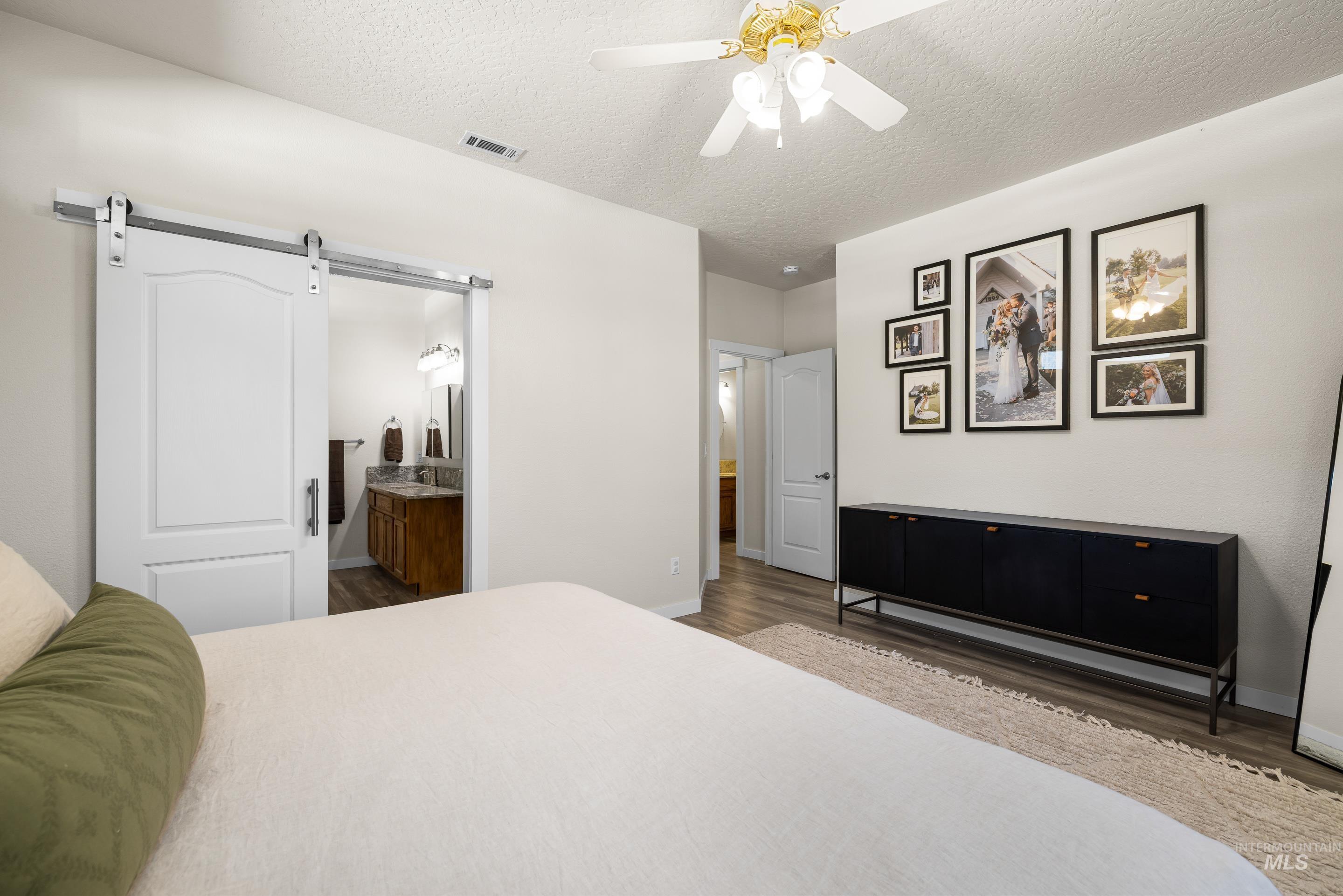 Bedroom featuring a textured ceiling, a barn door, wood finished floors, ceiling fan, and ensuite bath