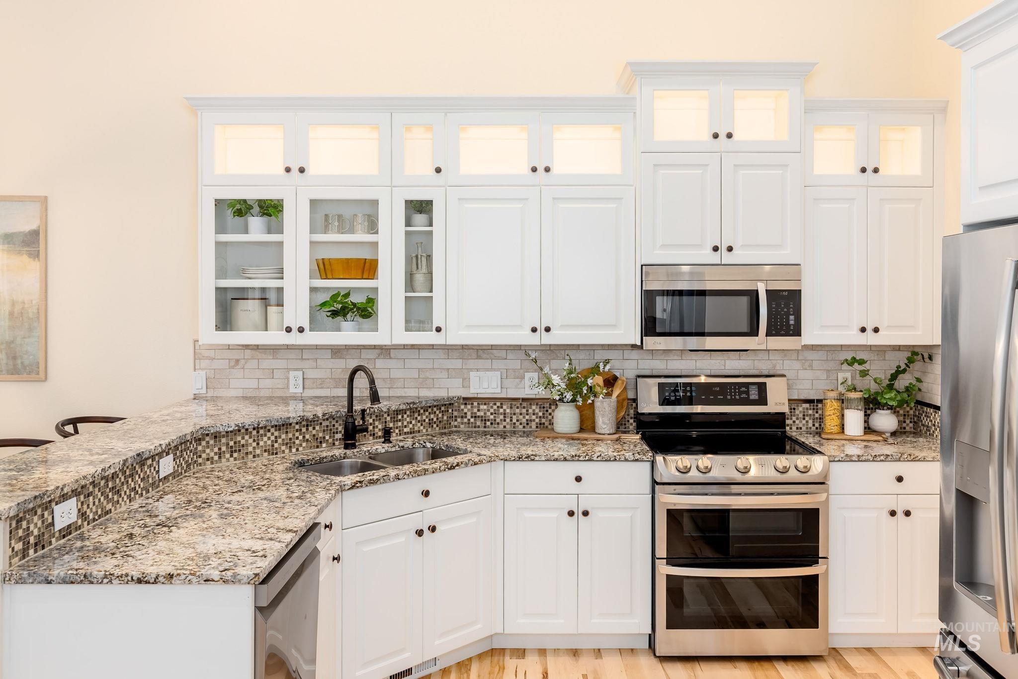 Kitchen with appliances with stainless steel finishes, white cabinets, light stone countertops, tasteful backsplash, and glass insert cabinets