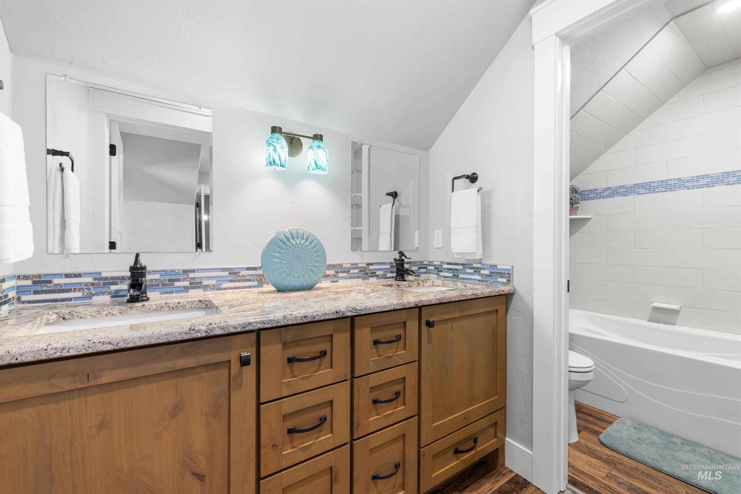 Bathroom featuring double vanity, dark wood-style floors, lofted ceiling, shower / bathtub combination, and tasteful backsplash