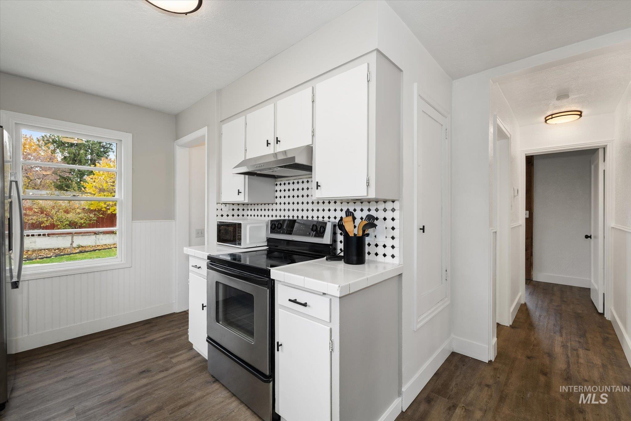Kitchen with stainless steel appliances, white cabinets, decorative backsplash, dark wood-style floors, and under cabinet range hood