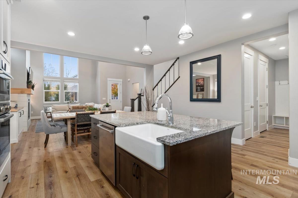 Kitchen with dark brown cabinets, light stone countertops, an island with sink, pendant lighting, and light wood-style floors