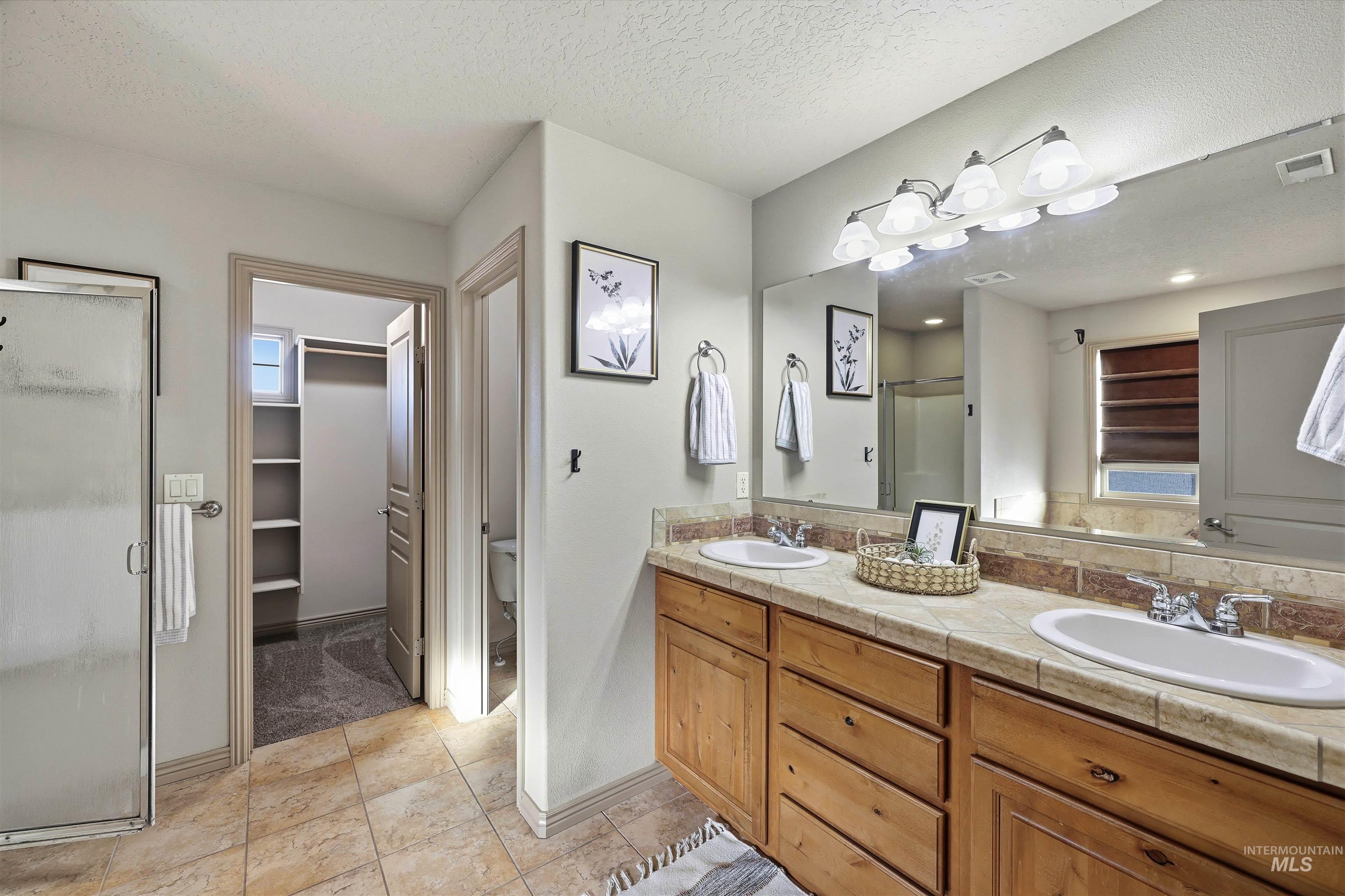 Bathroom featuring a shower stall, double vanity, a walk in closet, a textured ceiling, and light tile patterned floors